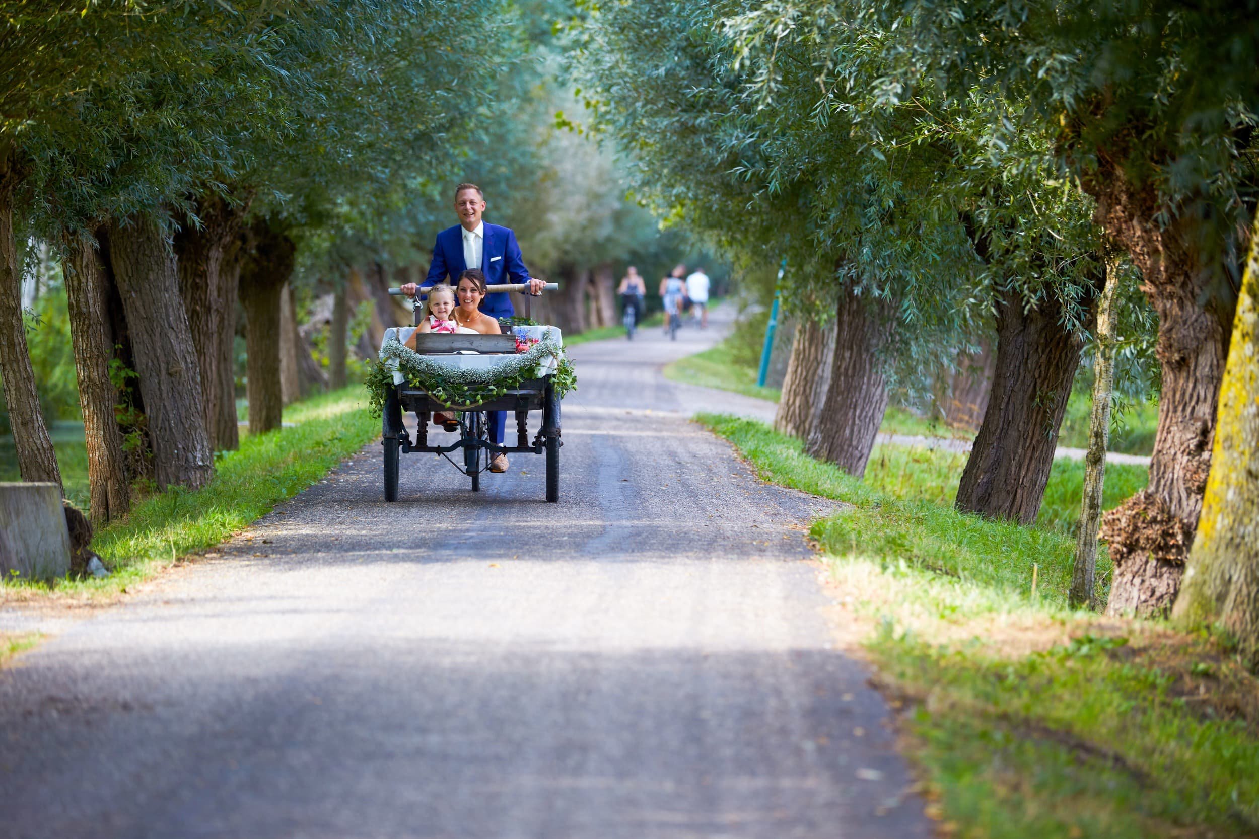 Gezicht op een man, vrouw en kind in een koets op een weg tussen bomen in een parkachtige omgeving.