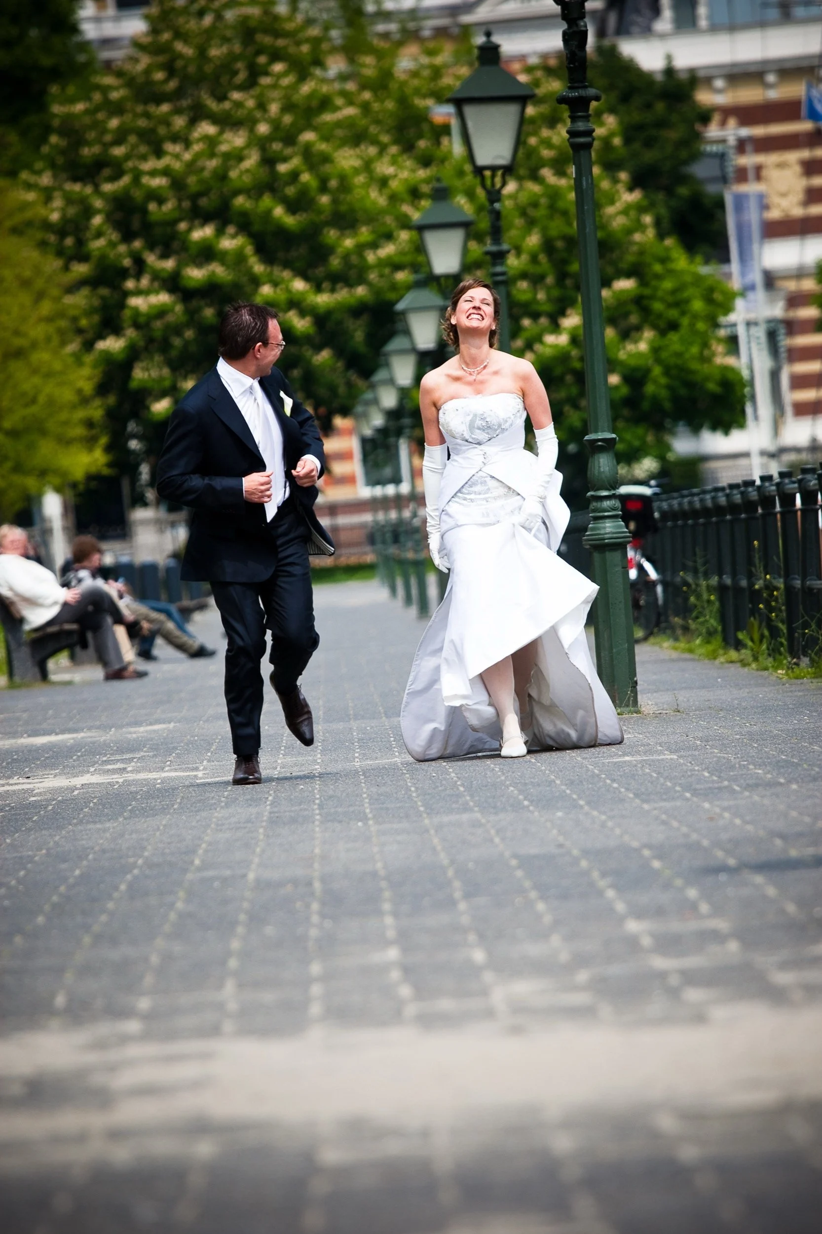 Trouwende echtpaar loopt lachend door een park terwijl ze elkaar aankijken, met groene bomen en straatlantaarns op de achtergrond.