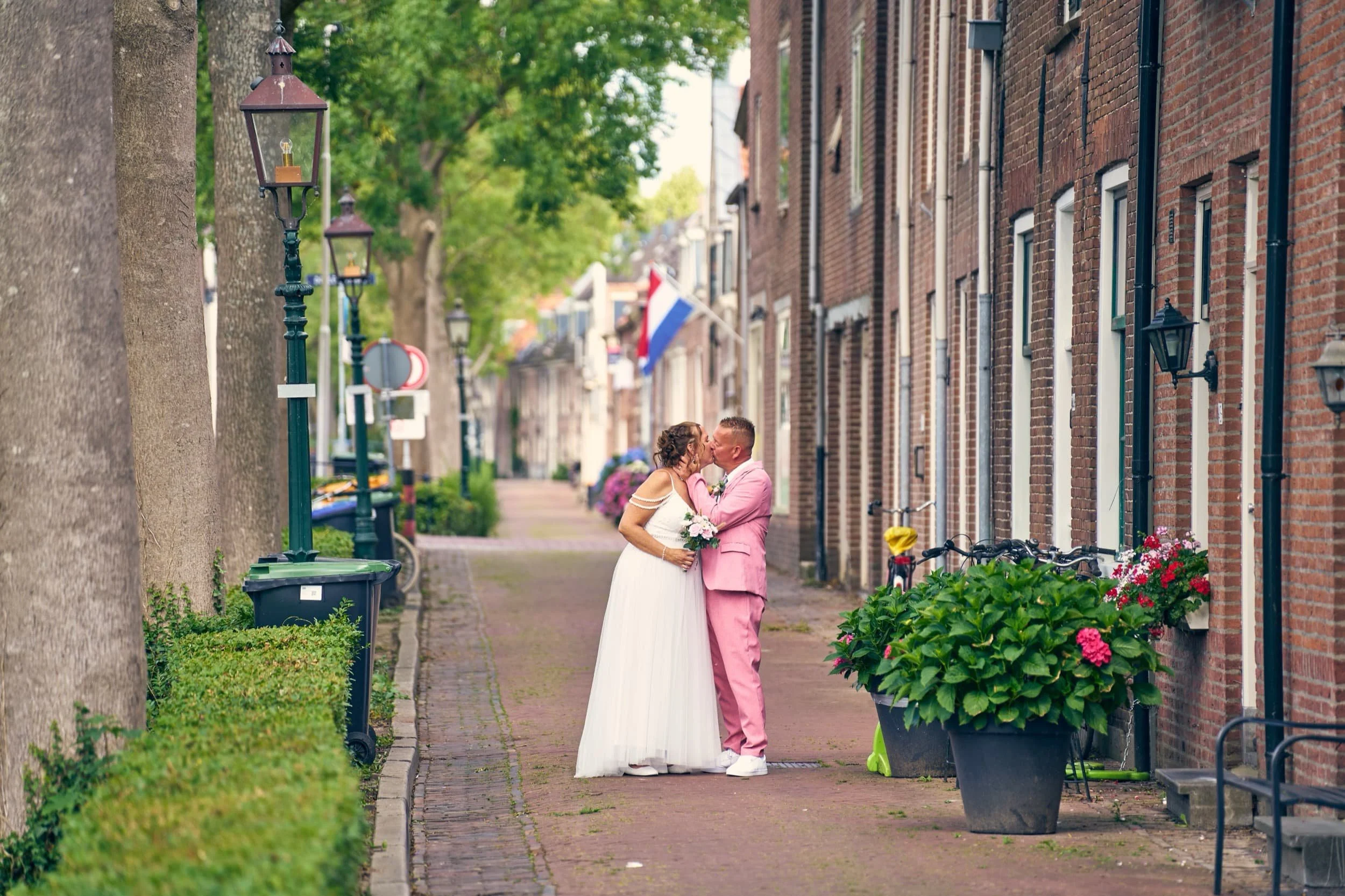 Een bruid en bruidegom die in een straat tegen elkaar aan staan, zoenend. De straat is weg van verkeer en heeft buizen, planten, fietsen en straatverlichting.