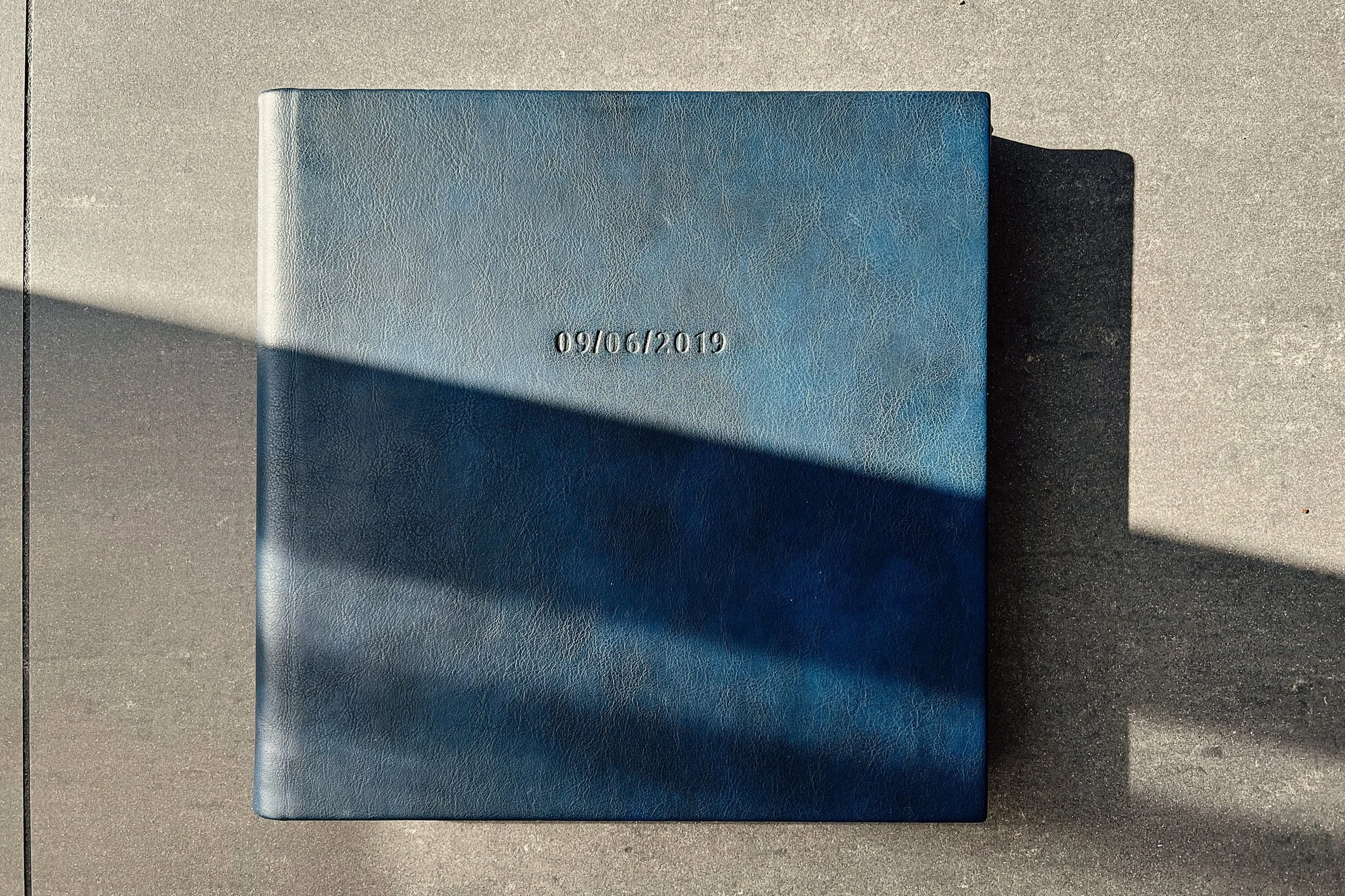 Blauwe leren boek met een datum '09/06/2019' in reliëf op de voorkant, gelegd op een betonnen oppervlak met schaduw.