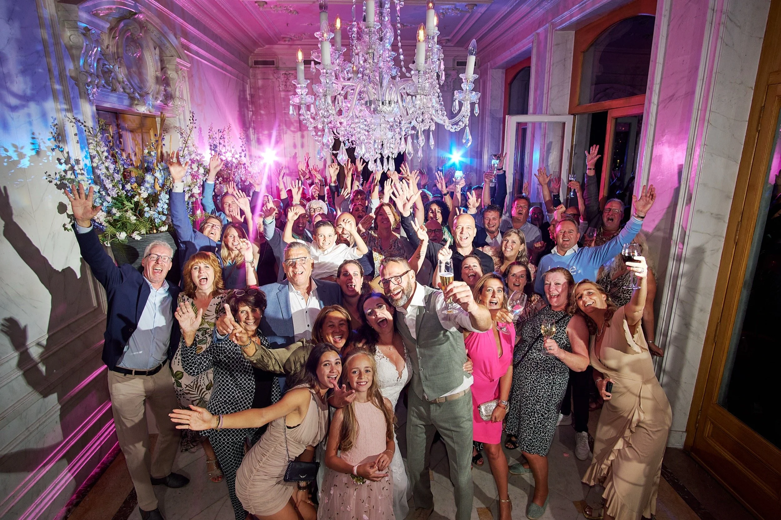 Een feestelijke groep mensen die lachen en plezier hebben in een elegante zaal met een grote kristallen chandelier, kleurrijke verlichting, bloemen en open ramen.