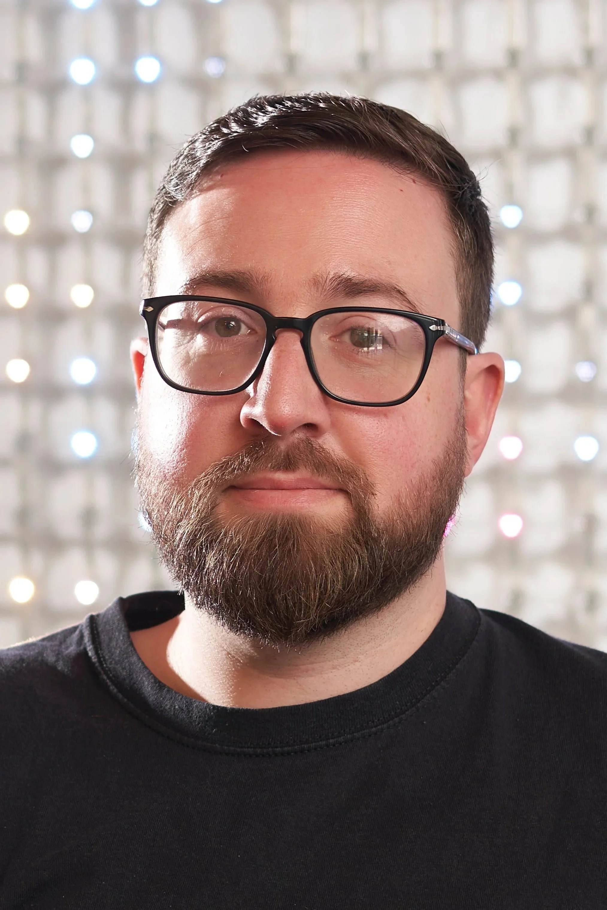 A close-up of a person's face, showing the upper part including hair, glasses, and beard. The background is a pattern of blurred lights.