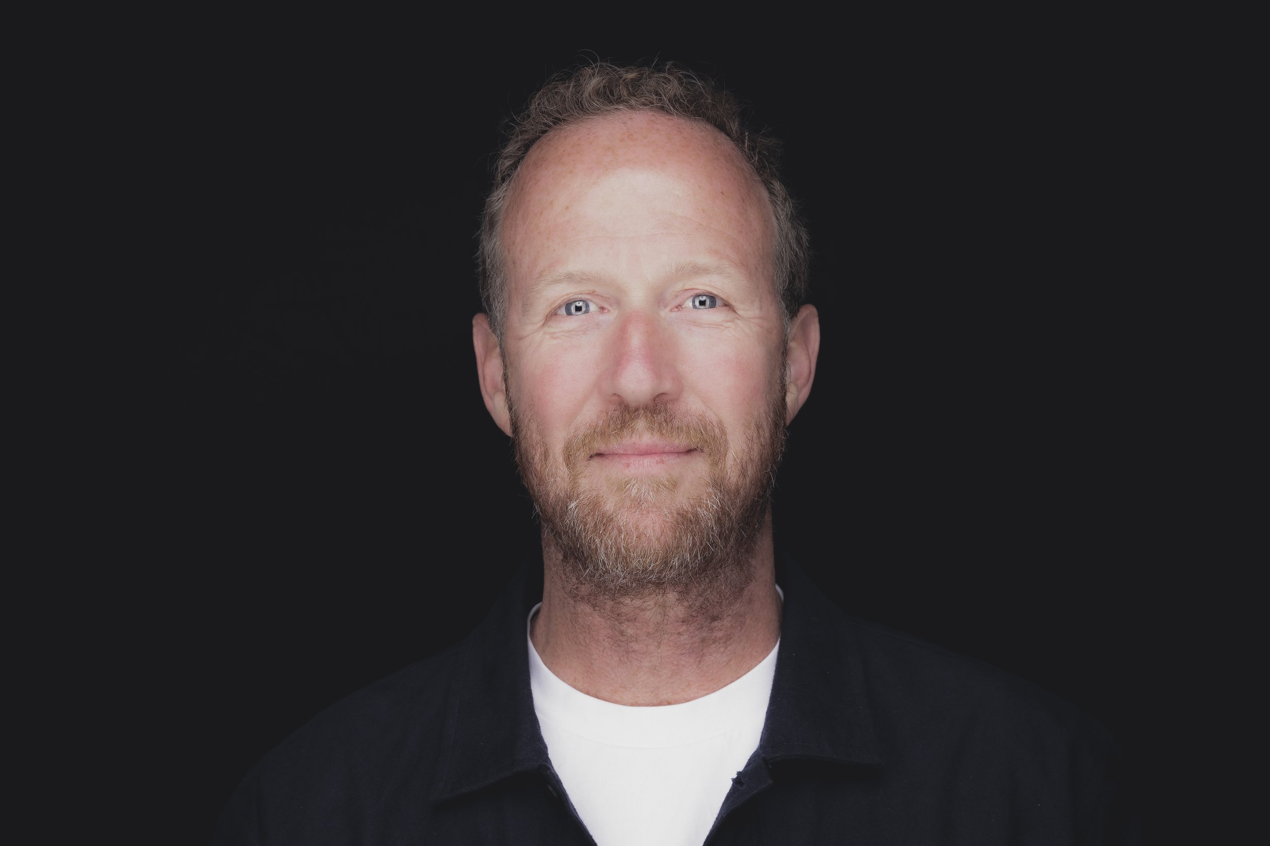 Portrait of a middle-aged man with glasses, a beard, and gray hair, wearing a blue sweater, standing indoors with blurred windows in the background.