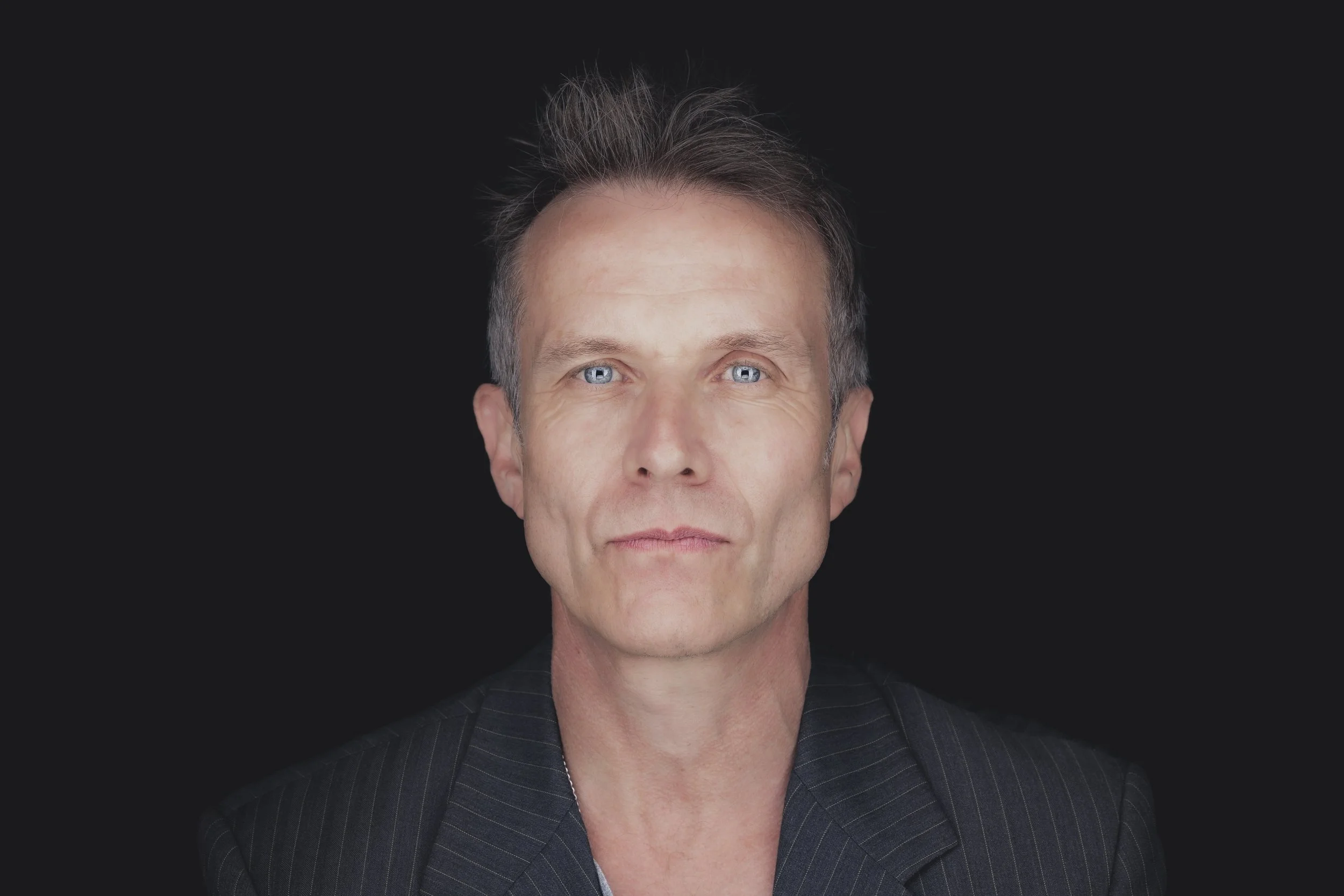 A middle-aged man with light skin, blue eyes, and gray hair, wearing a dark striped suit jacket, against a black background.