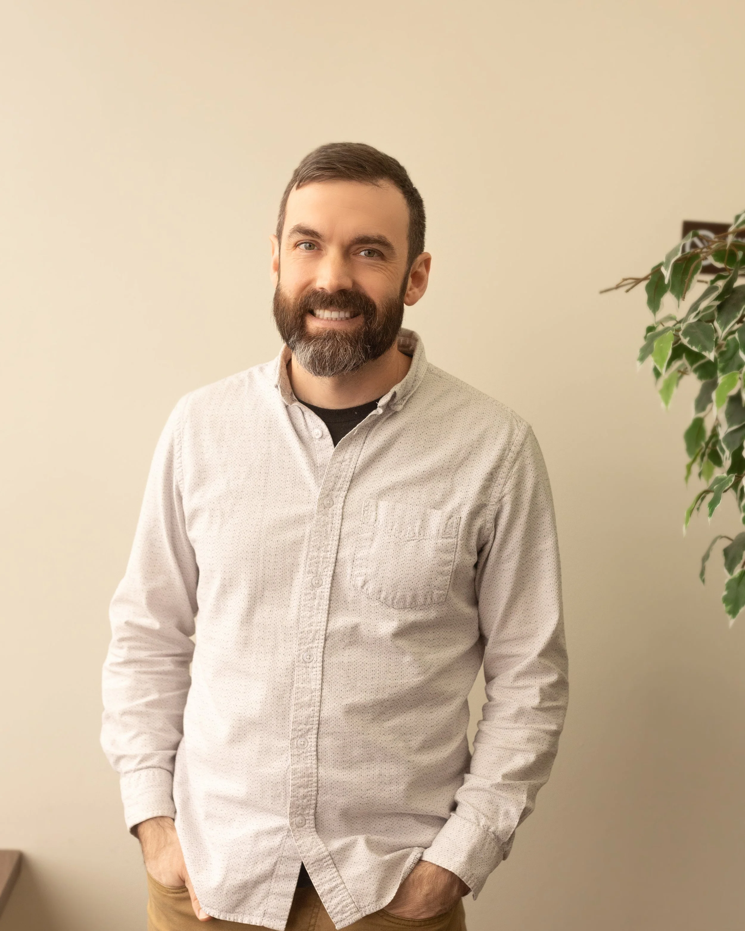 A smiling man with a beard and short dark hair, wearing a light-colored button-up shirt, standing indoors in front of a plain beige wall and a leafy green plant.