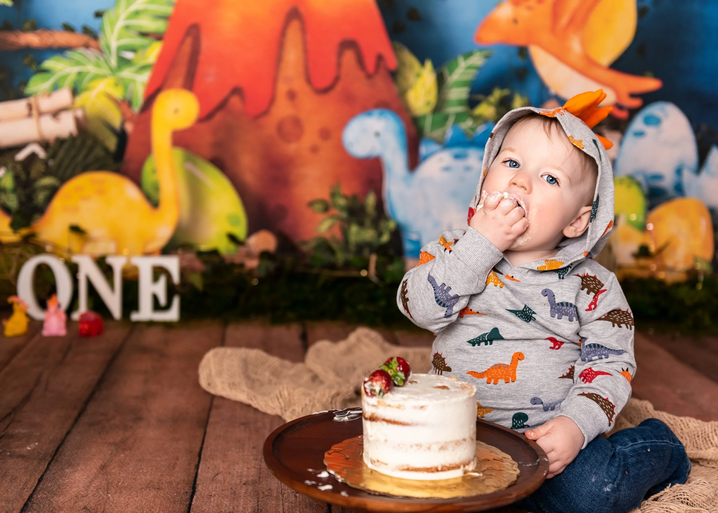 picture of little baby boy in dino hoodie eating a cake at his one year cake smash photography shoot in a studio in Canton, GA