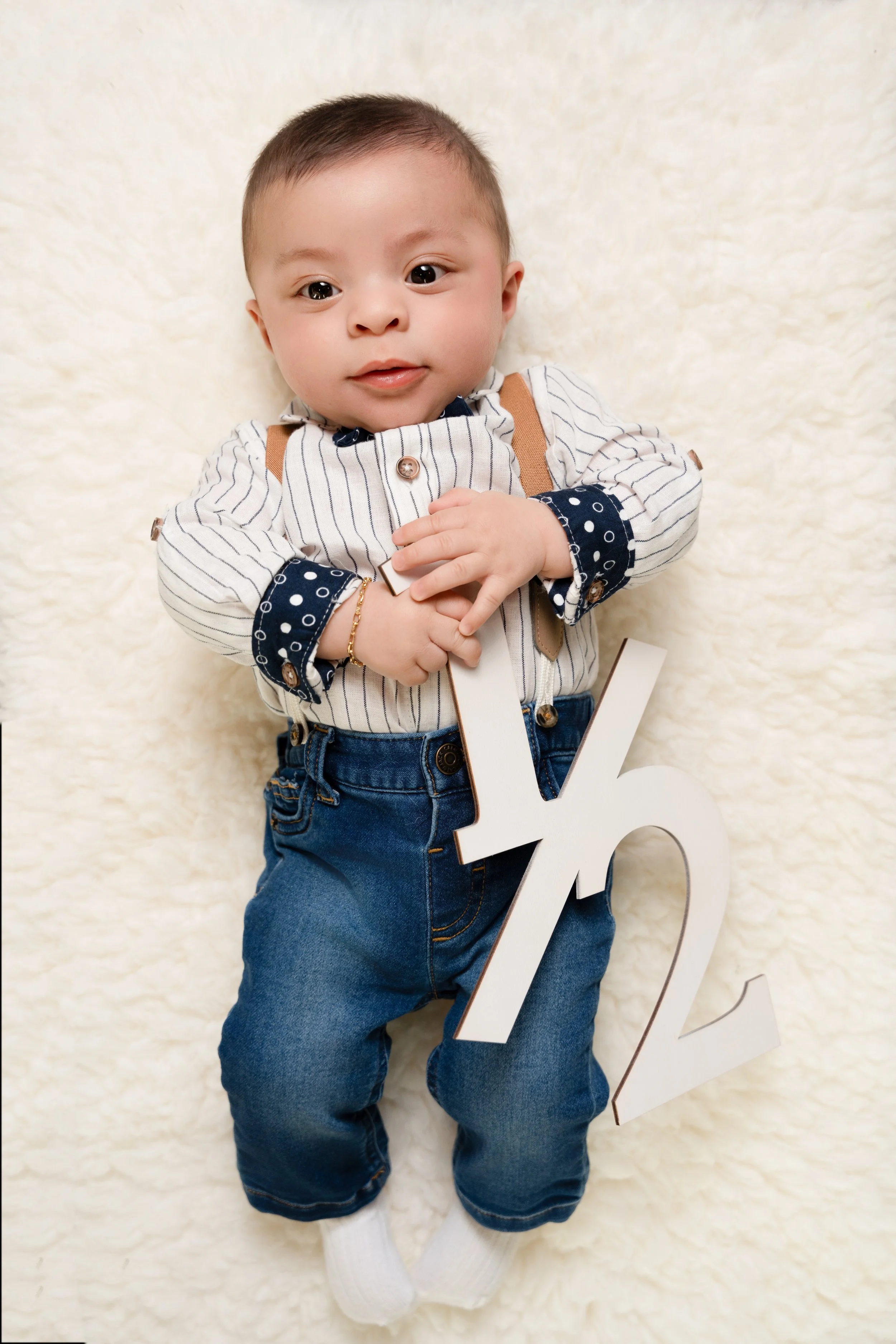 picture of baby boy in outfit with suspenders laying on white fur holding a wooden 1/2 to commemorate his half birthday