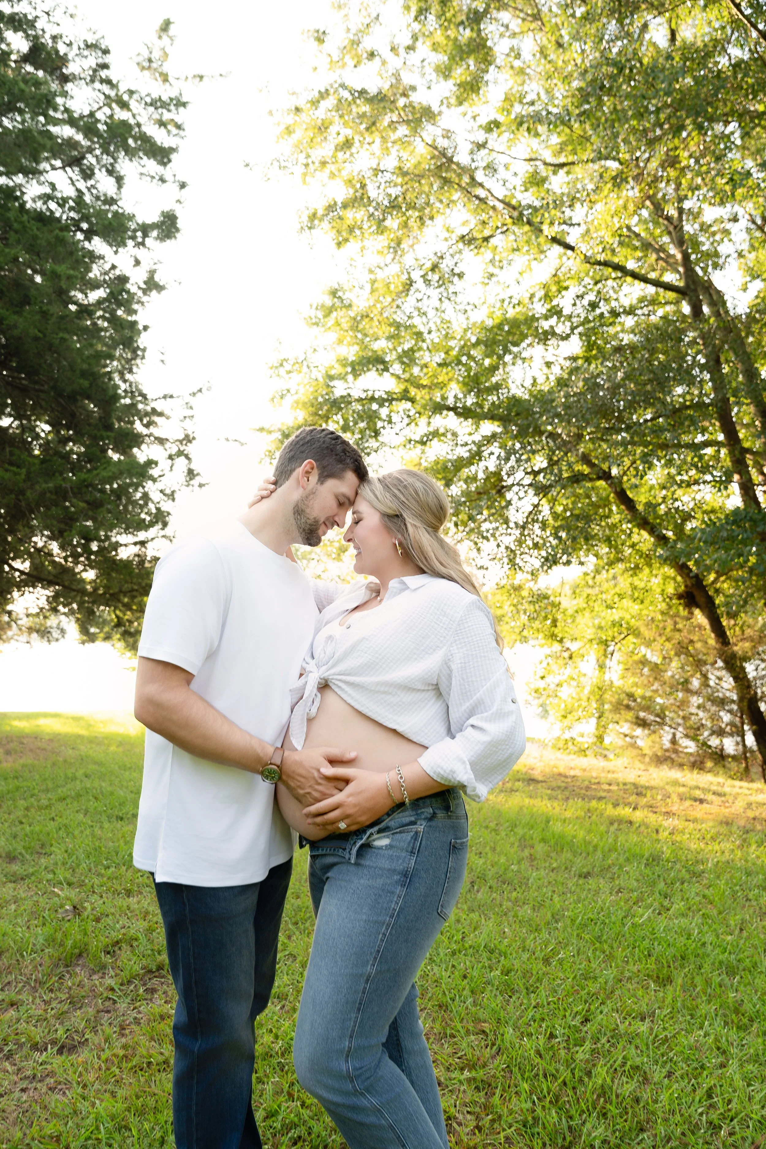 A couple standing outdoors on a grassy field, with the woman pregnant, gently touching her belly and holding hands, their foreheads touching, surrounded by lush green trees with sunlight filtering through.