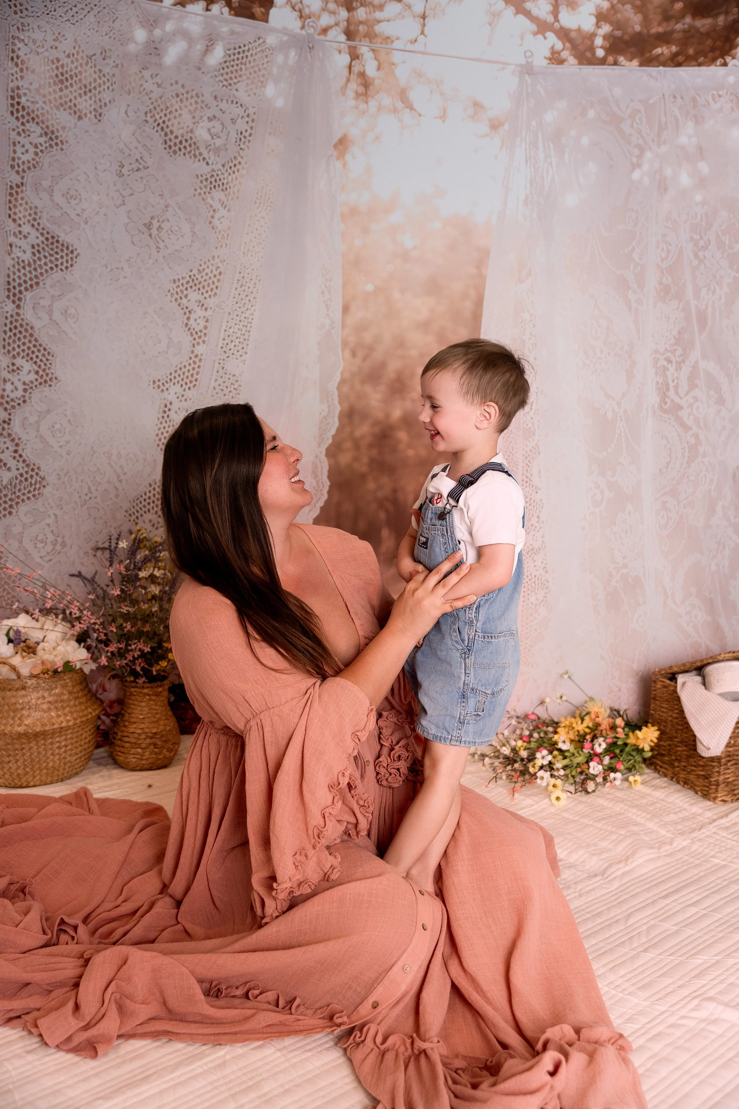 image of mom in gorgeous pink dress with little boy in overalls smiling at each other at a sunset clothesline set in a photography studio in Canton, GA
