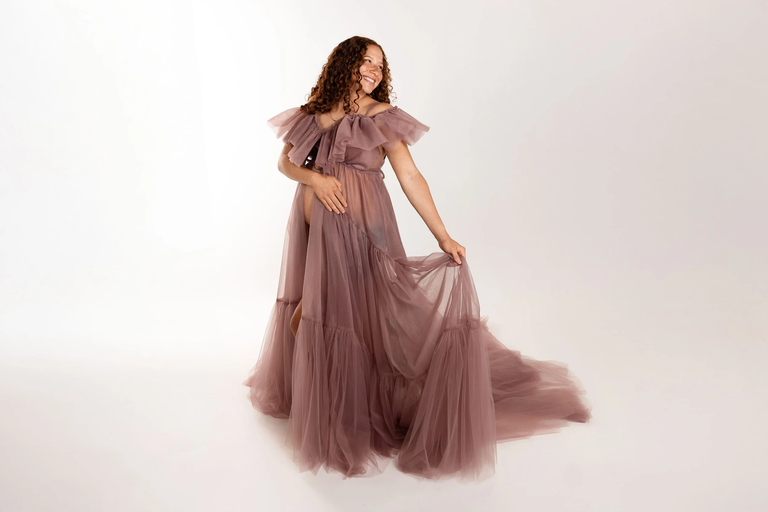 A woman with curly hair wearing a mauve-colored tulle gown with ruffled shoulders, smiling and holding the skirt of her dress.