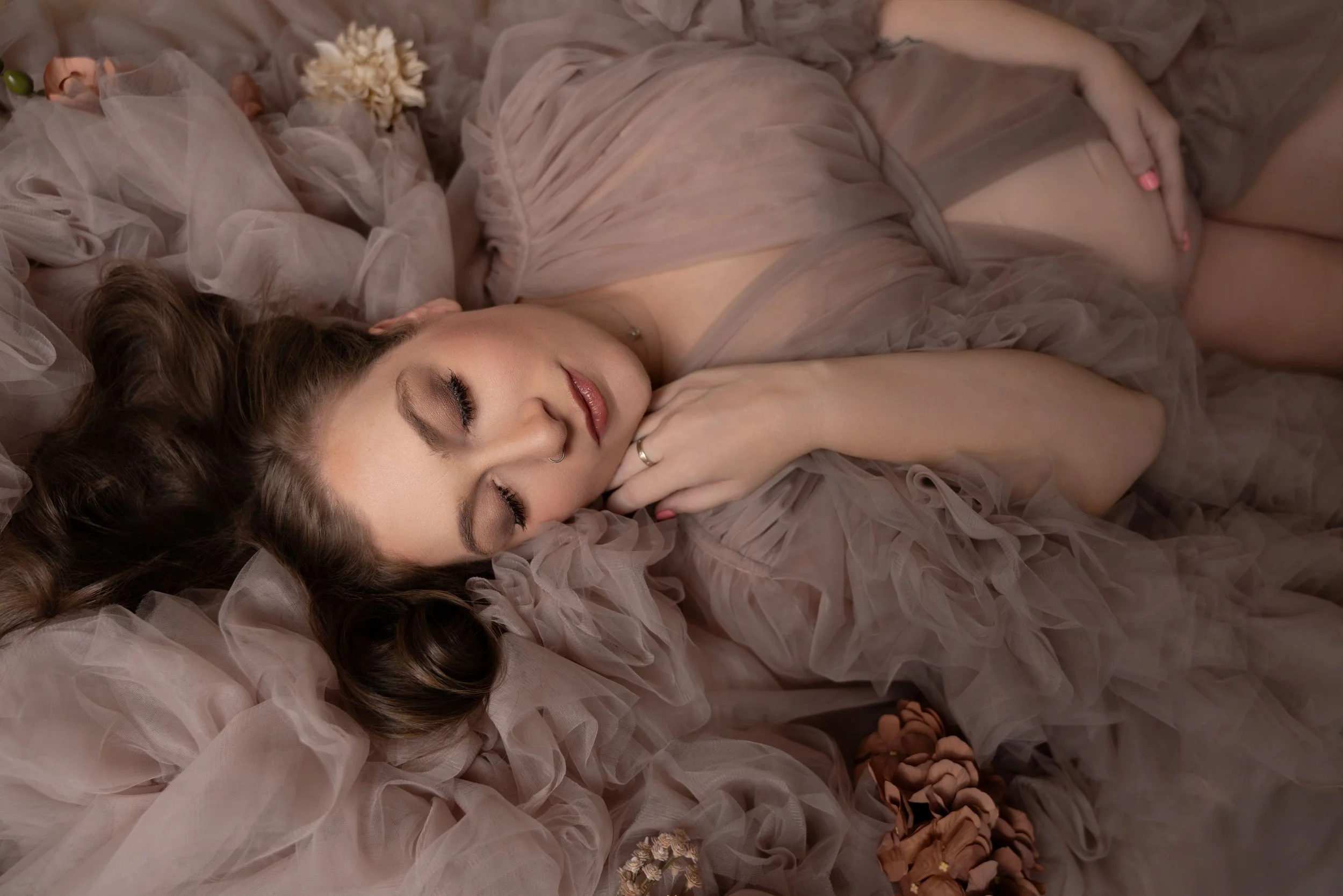 this image is showing a beautiful mom-to-be laying down on her side in a beautiful tulle light pink gown showing her baby belly and surrounded by flowers