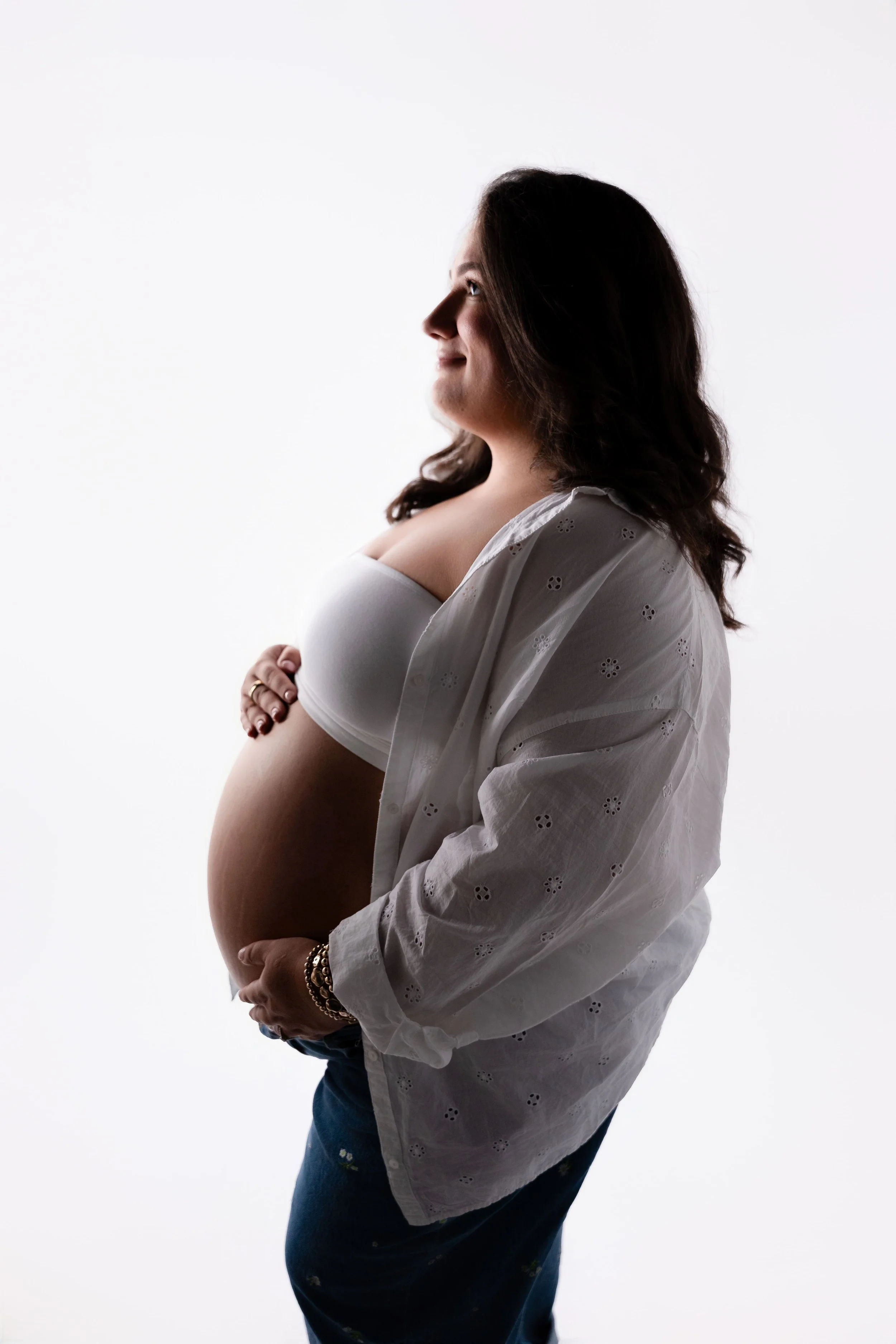 Side profile of a pregnant woman with dark hair, smiling, wearing a white off-the-shoulder top and a white eyelet shirt, standing against a white background.