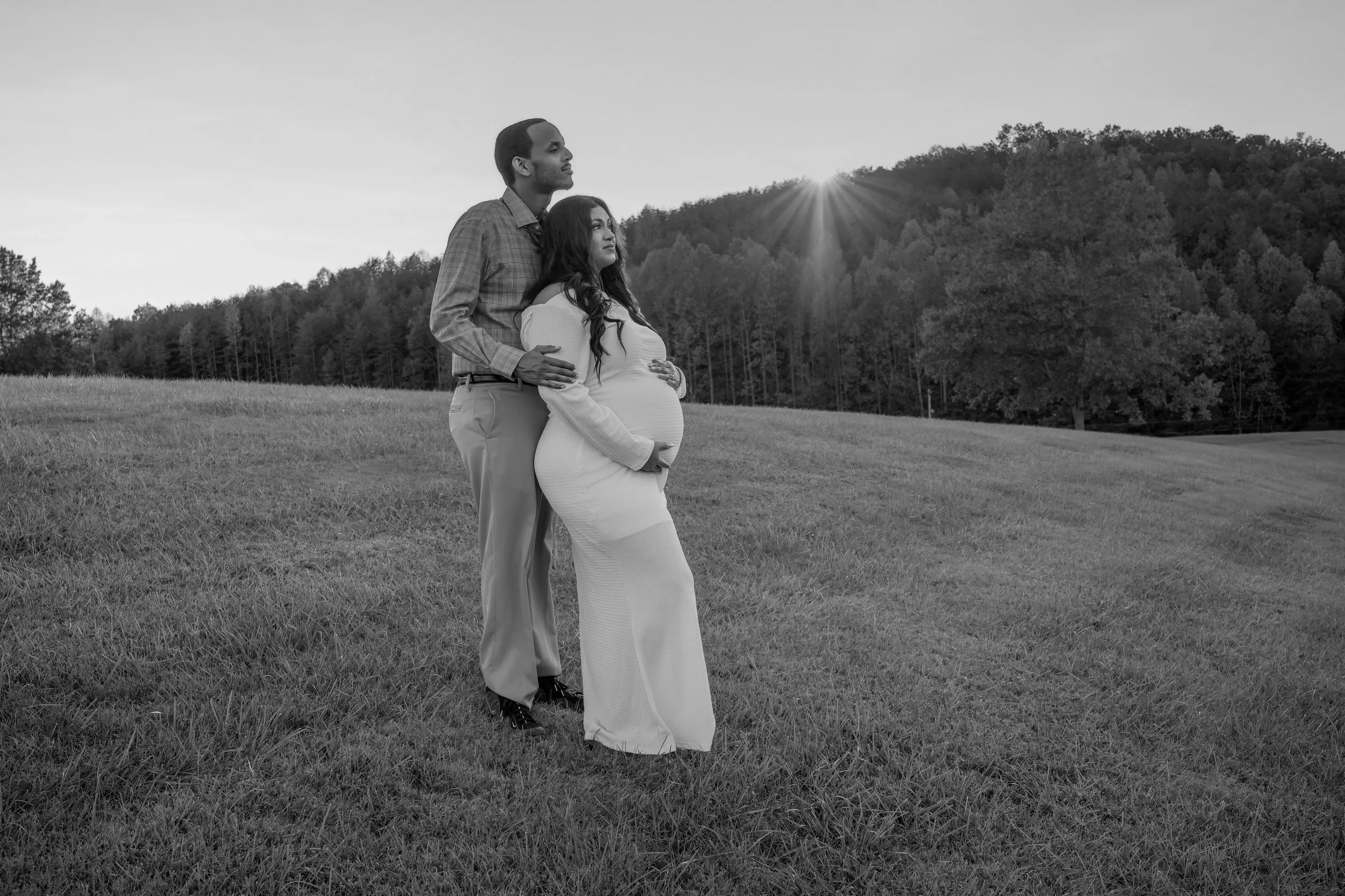 Pregnant woman smiling while holding her belly, wearing an elegant maternity dress with the North Georgia mountains behind her with her partner