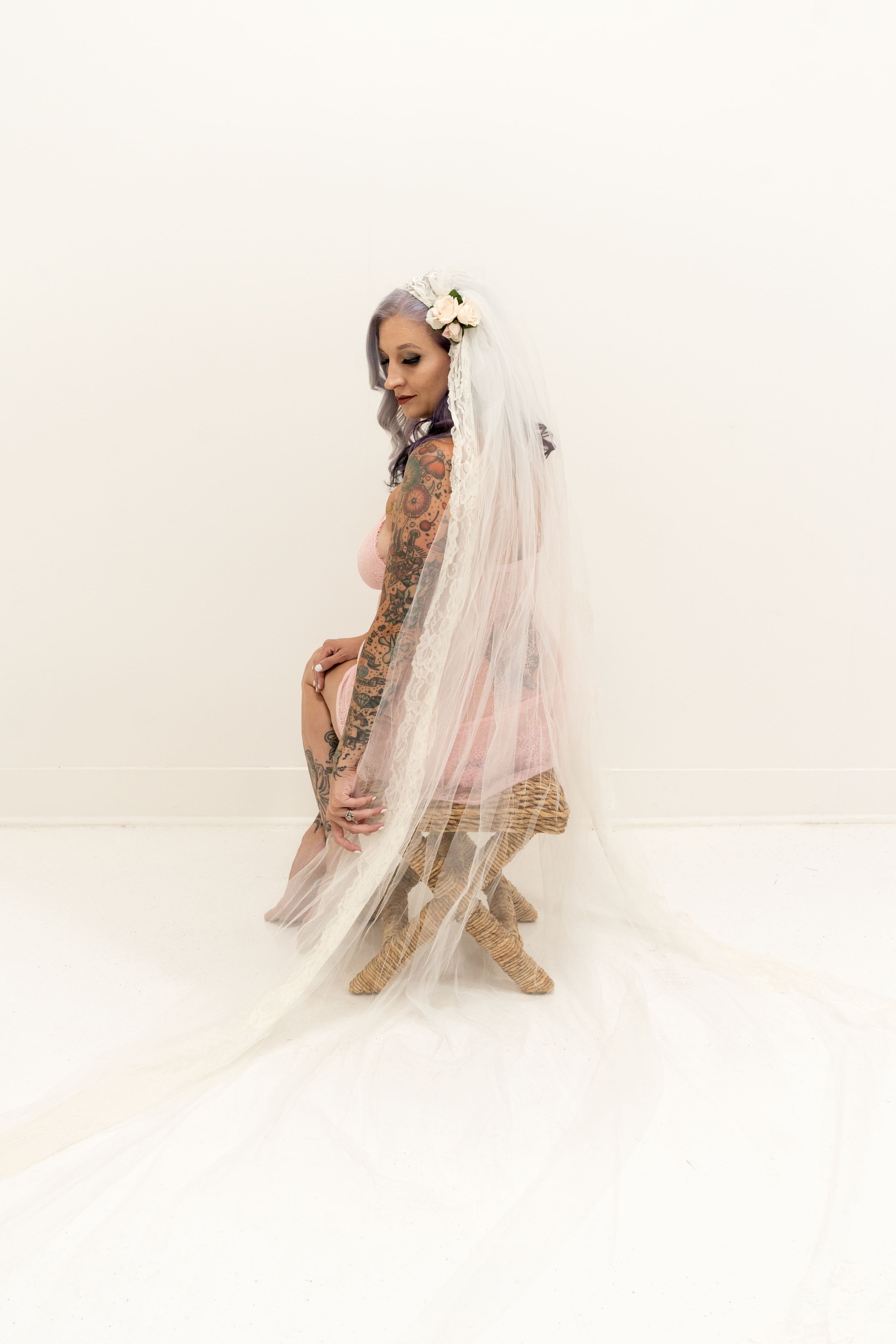 A woman with purple hair, tattoos on her arms, and in a pink dress, wearing a white veil with flowers, kneeling on a small woven stool, in front of a plain white wall.