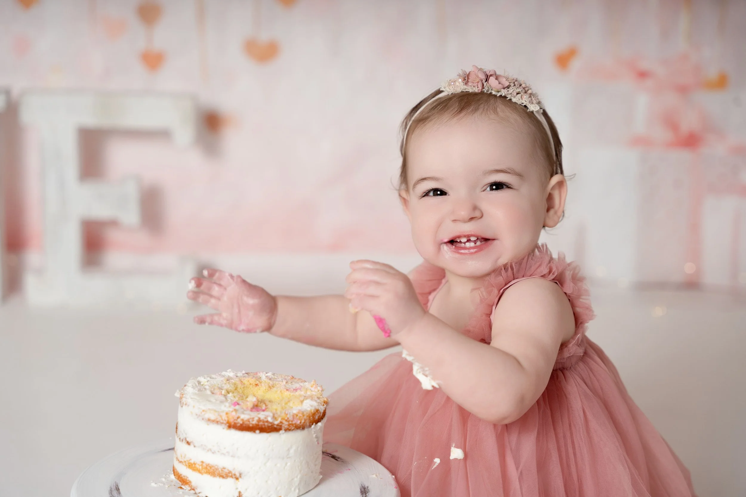 this image shows a happy baby girl dressed in a pink tulle dress and matching headband with white icing on her fingers and arms with a white cake on a cake stand in front of her and pink decor behind her celebrating her one year birthday