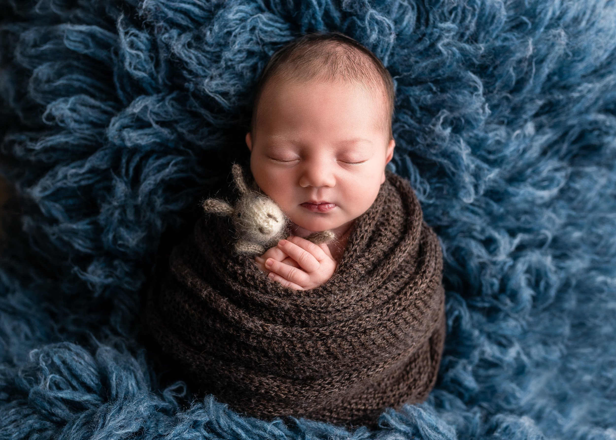 this image is of a sweet newborn baby wrapped snug in a brown newborn wrap sitting up and asleep holding a little giraffe lovey with a blue fur underneath baby