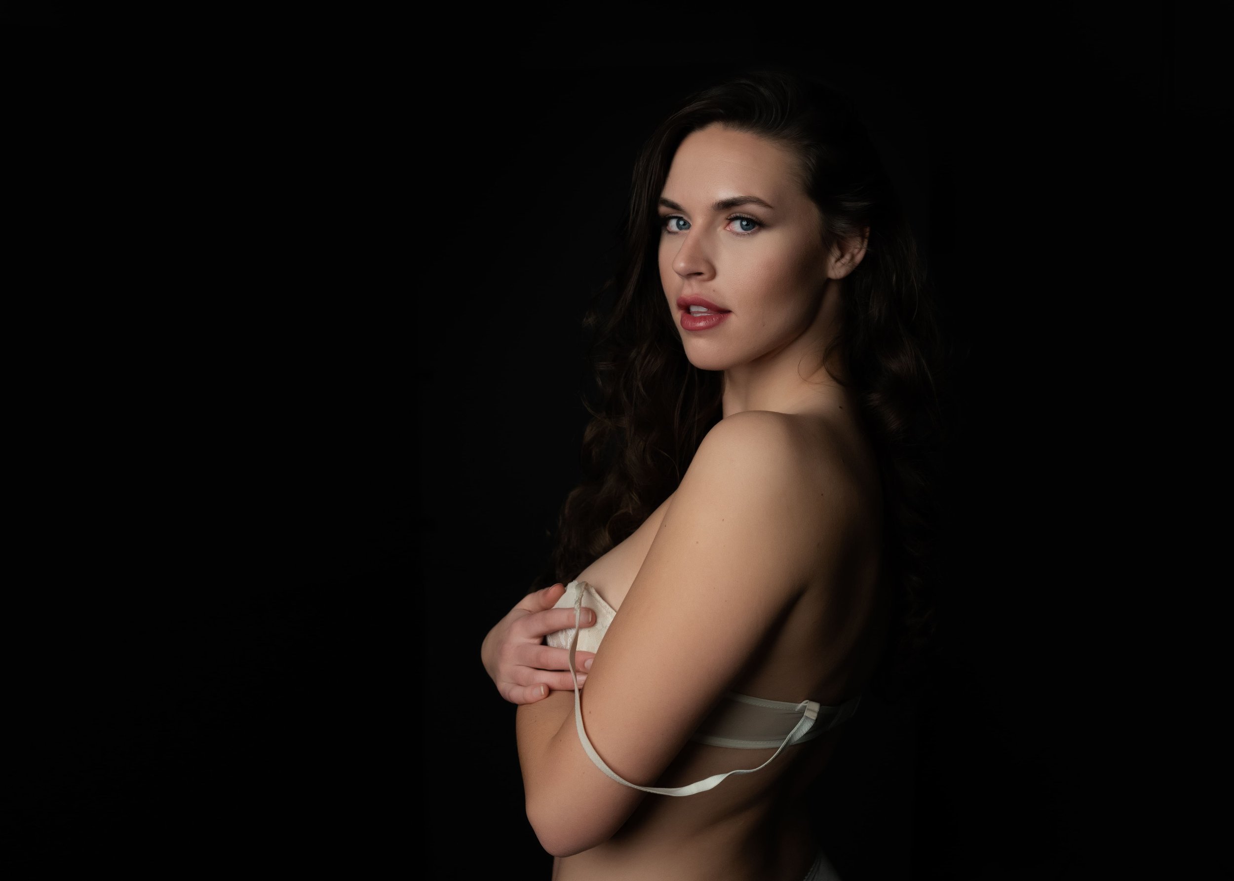 A woman with long dark hair and blue eyes posing topless against a black background, holding a piece of fabric across her chest.