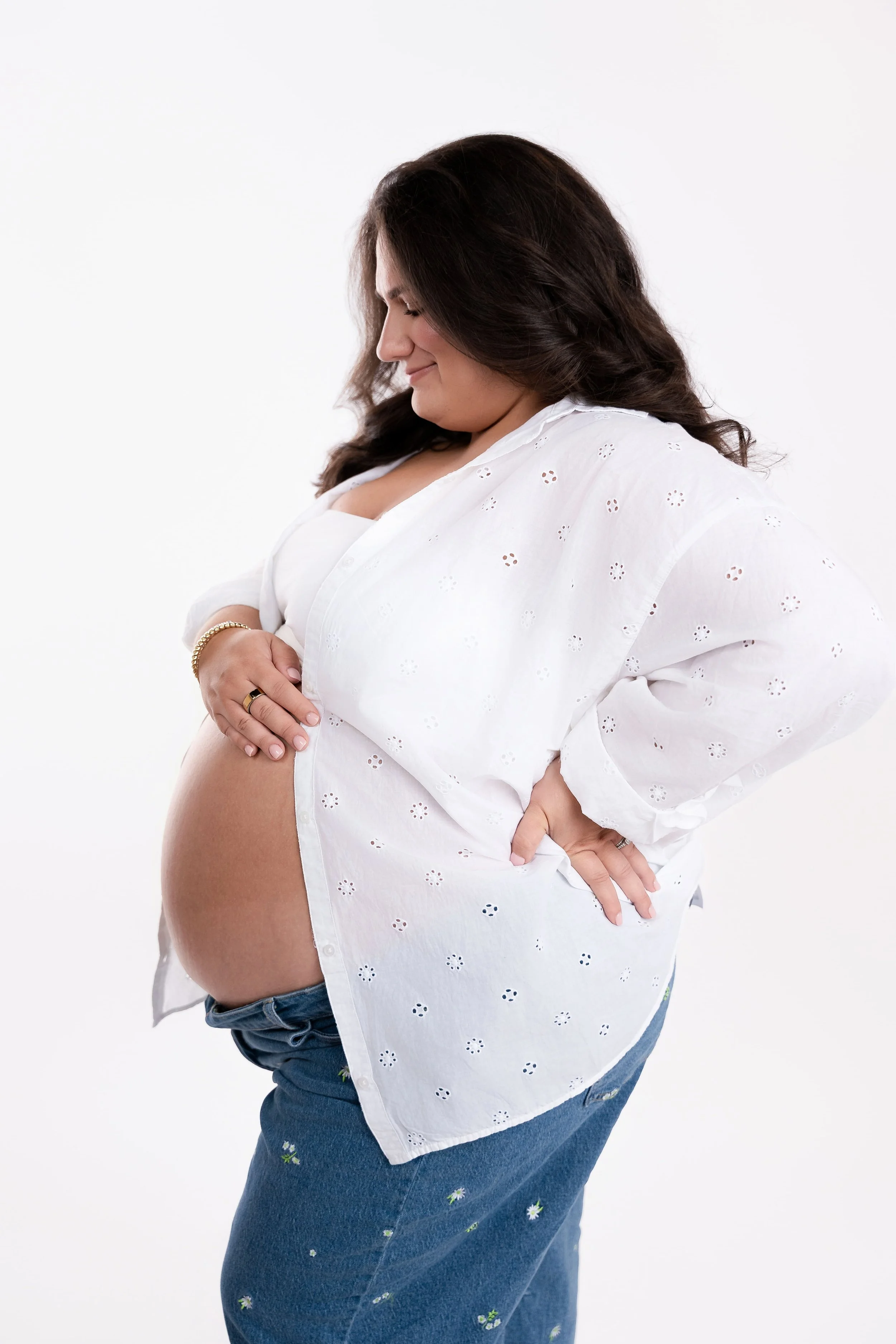 Pregnant woman with long dark hair wearing a white eyelet blouse and blue jeans, smiling and looking down at her baby bump, standing against a plain white background.