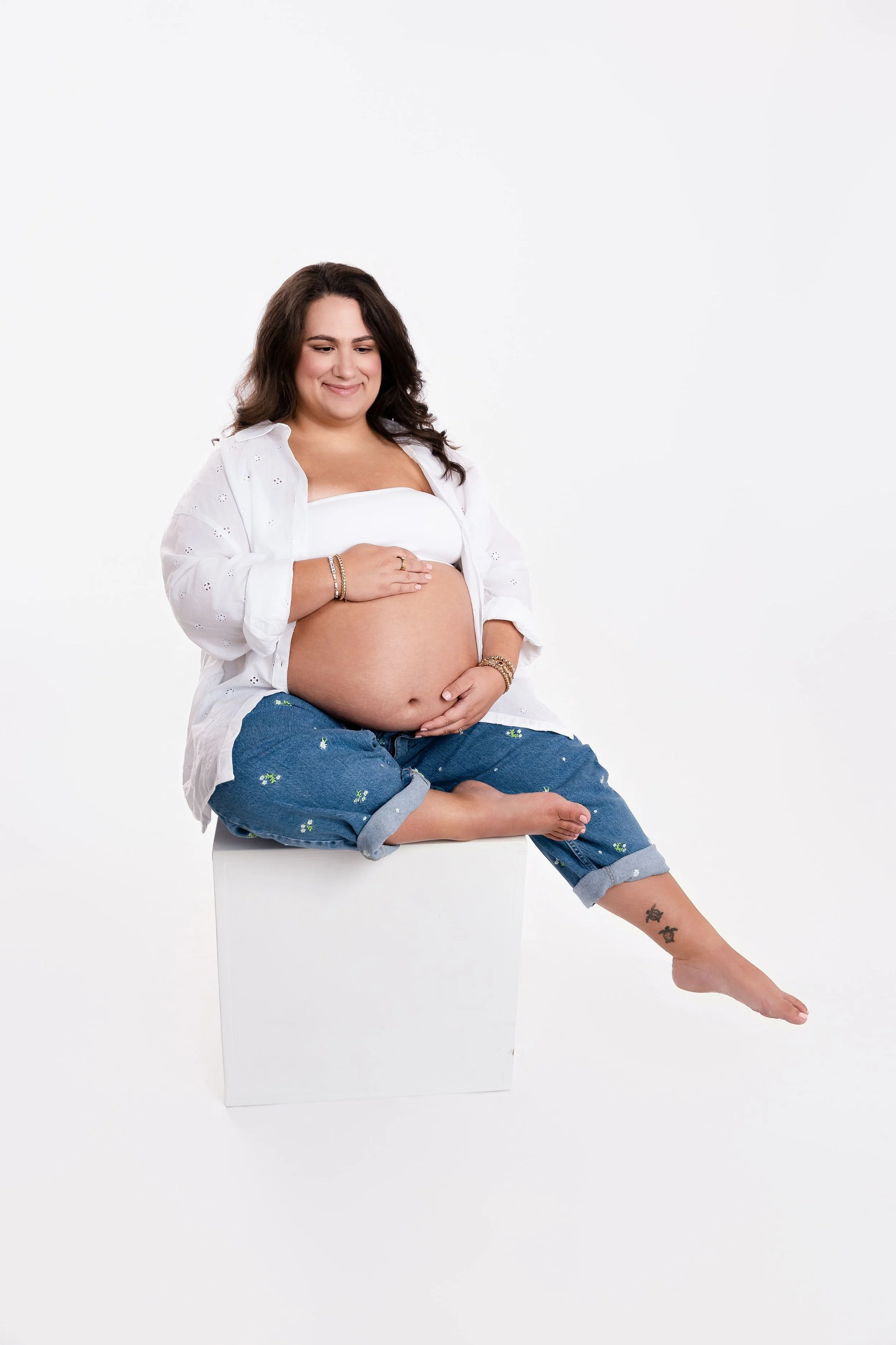 A pregnant woman sitting on a white block against a plain white background, smiling and looking down at her belly, wearing a white top, a partially unbuttoned white shirt, and blue jeans with floral embroidery, with tattoos on her left leg.