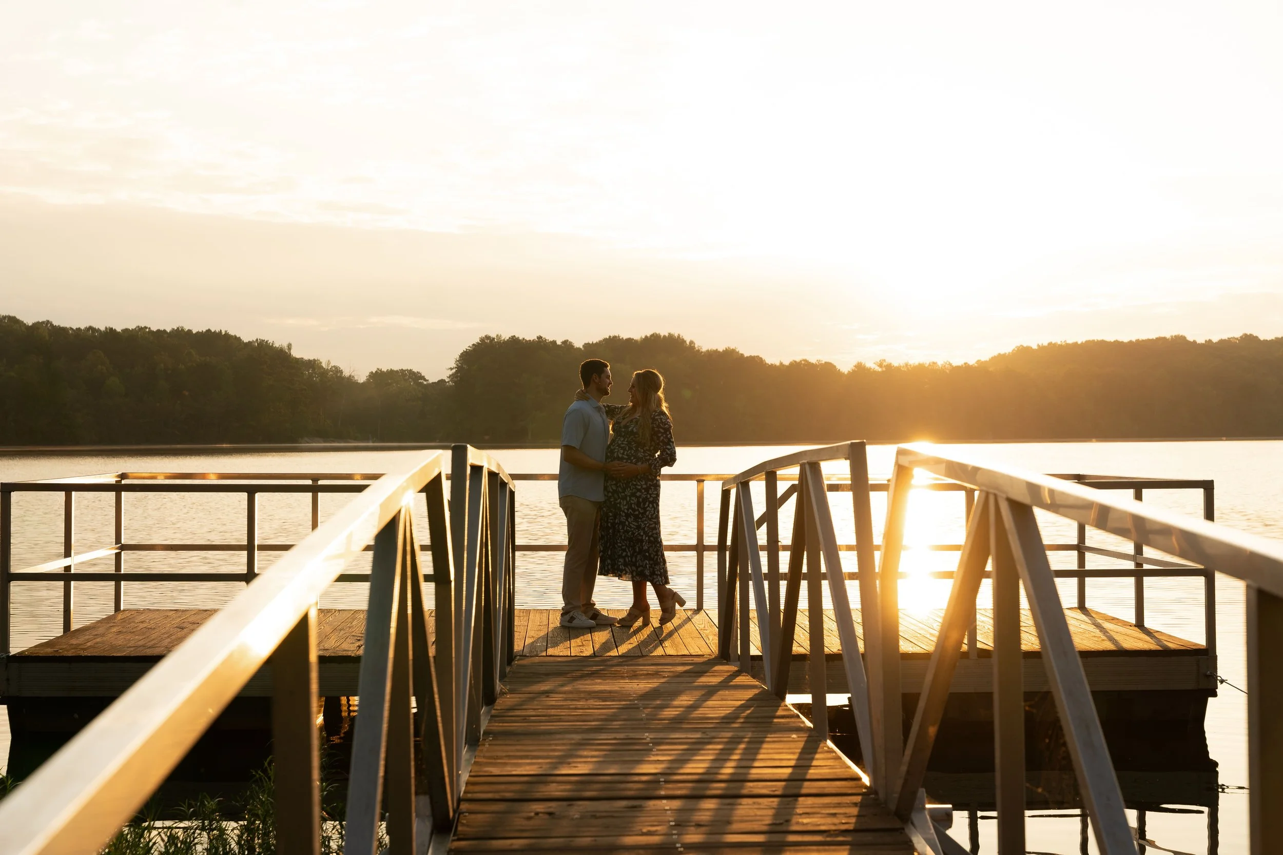 A couple stands on a wooden dock by a lake, holding hands and looking at each other during sunset, with trees in the background.