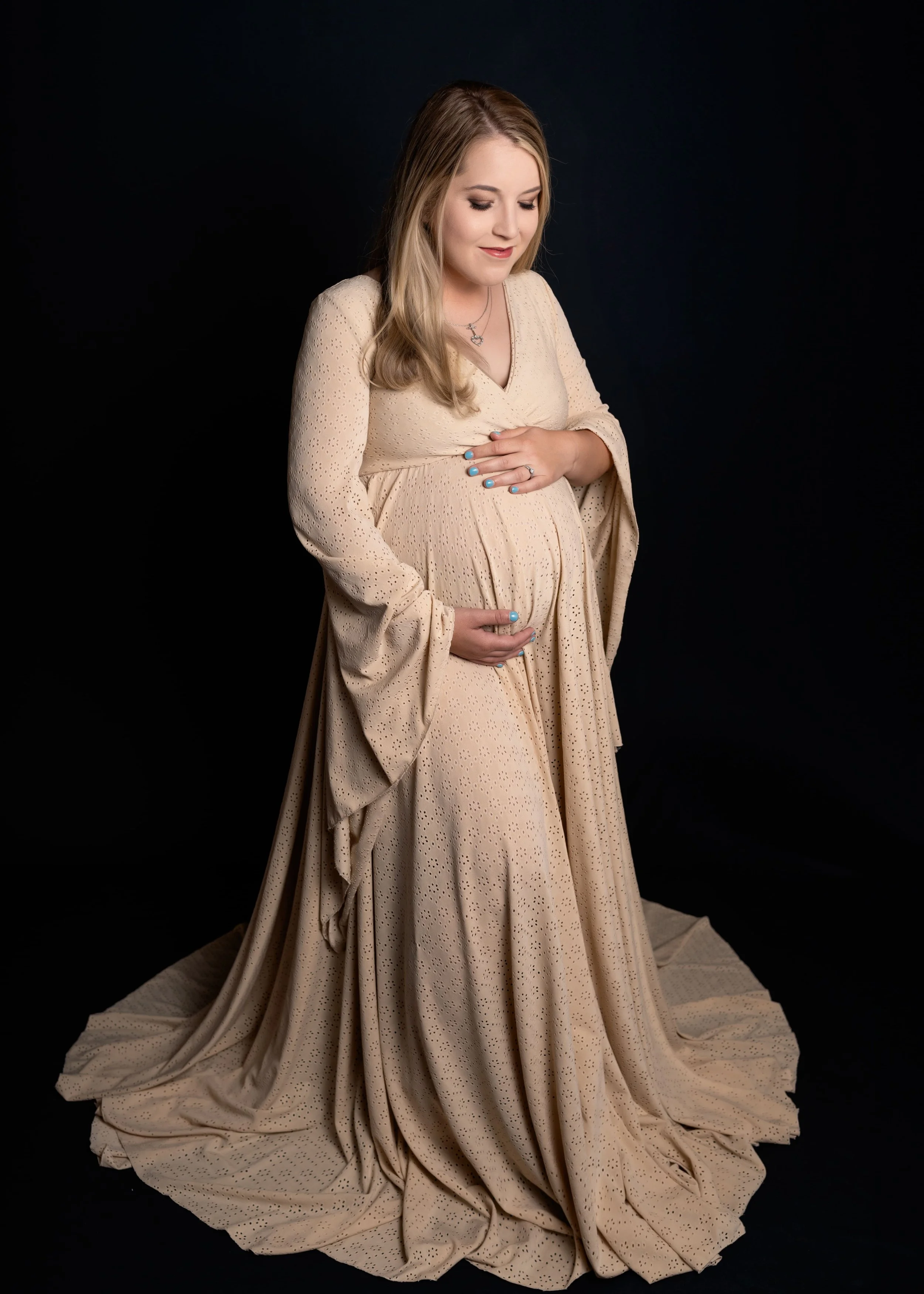 A pregnant woman in a flowing beige dress with eyelet details, gently holding her belly with both hands, standing against a black background.