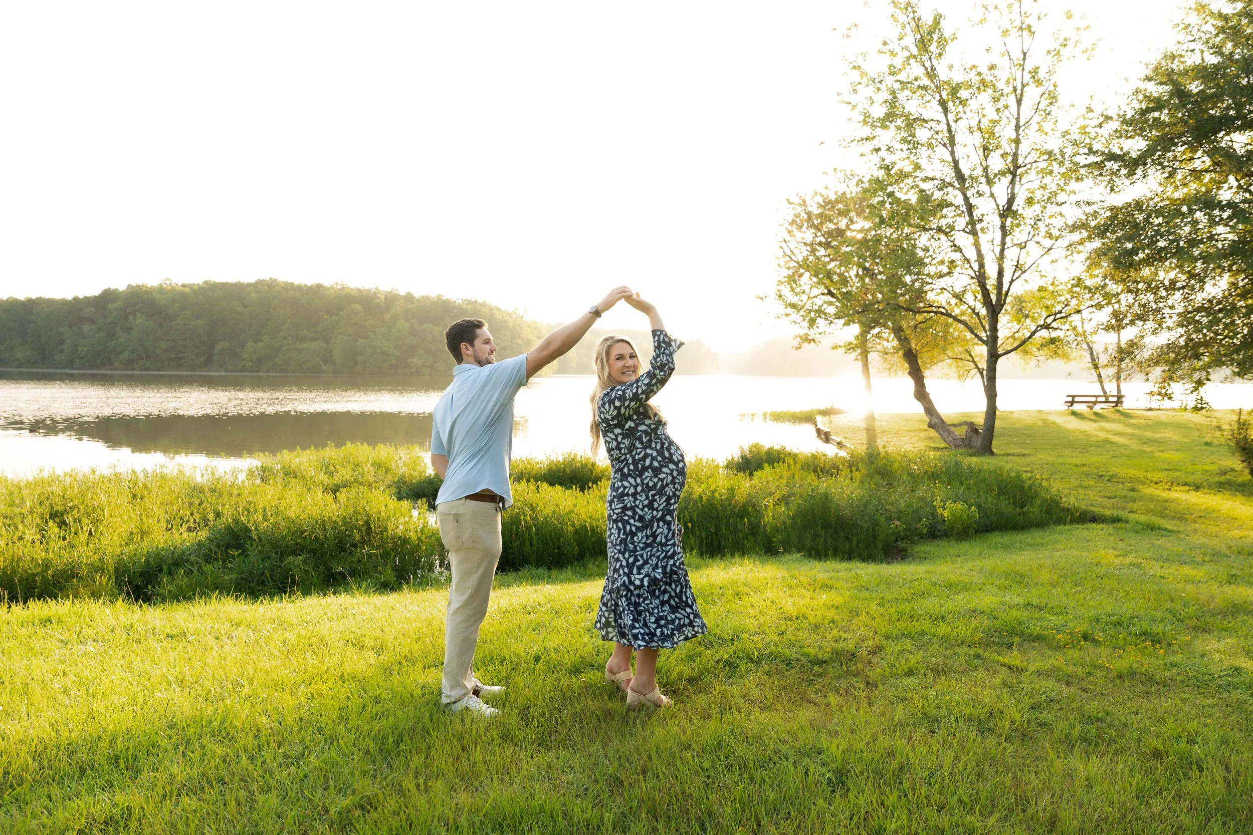 A couple dancing by a lakeside during sunset, with trees and picnic tables in the background.