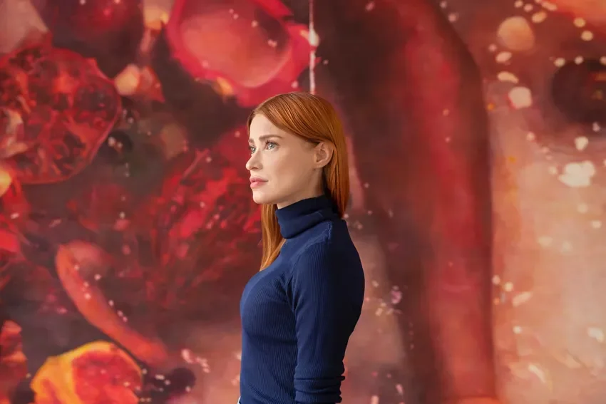 A woman with red hair wearing a dark blue turtleneck sweater, standing sideways in front of an abstract, colorful background.