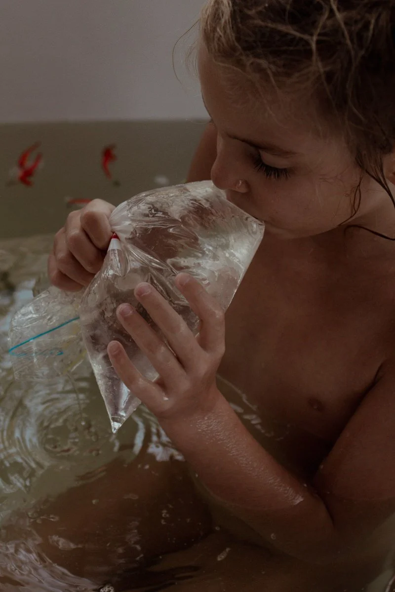 Junge Frau mit nassem Haar, die eine Plastiktüte mit Wasser in einer Badewanne hält und darin trinkt.