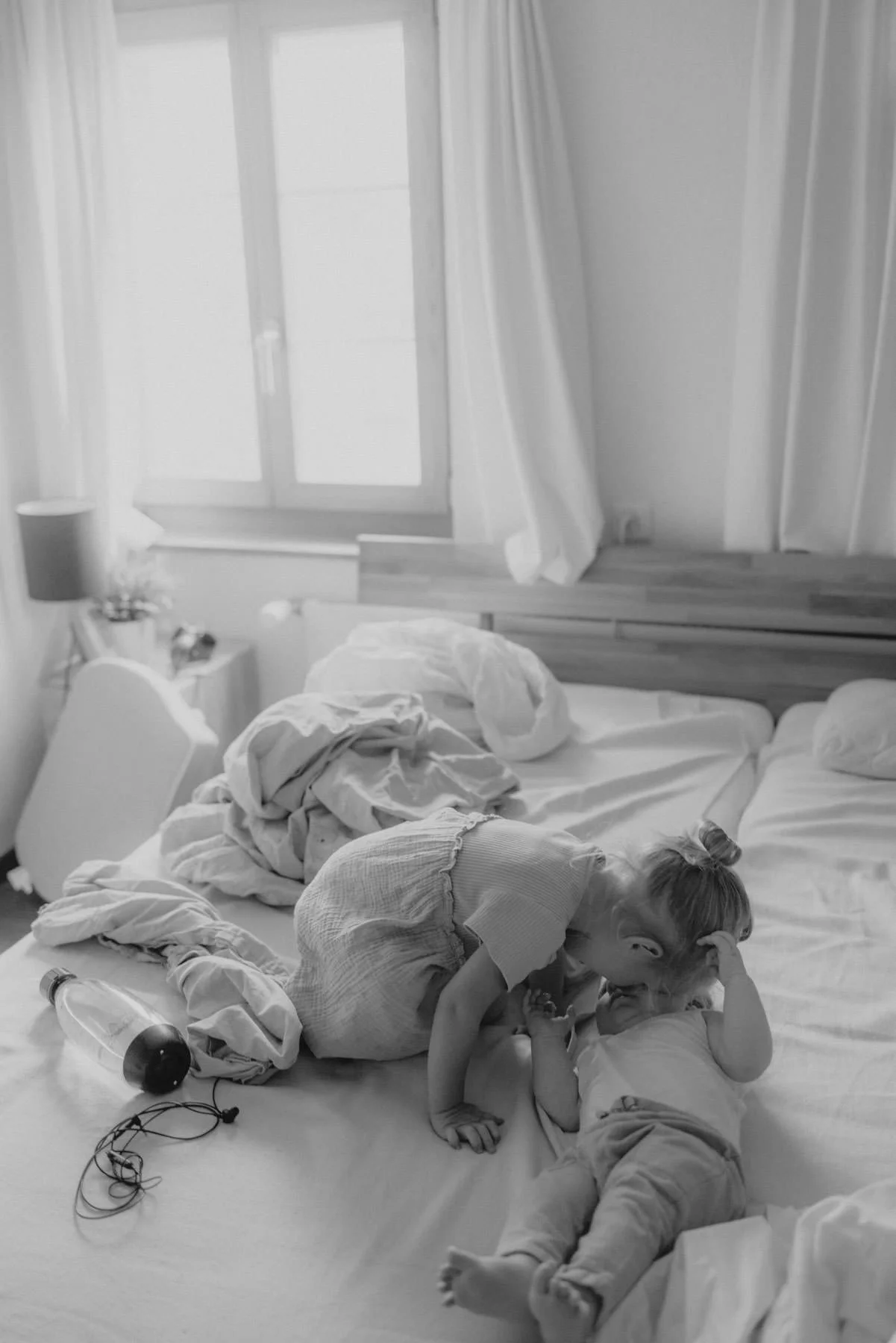 Ein Kind und ein Baby spielen auf einem unordentlichen Bett in einem Schlafzimmer mit einem Fenster im Hintergrund