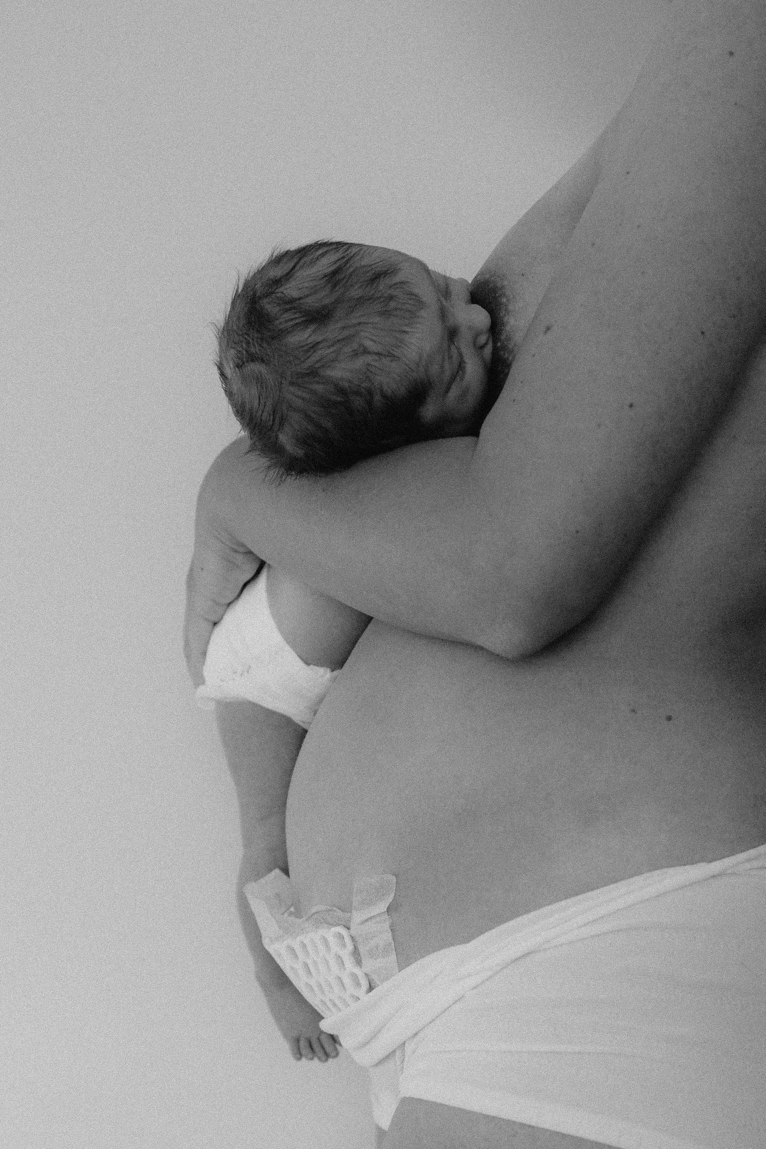 Ein neugeborenes Baby ruht auf dem Arm eines Erwachsenen, der nur teilweise im Bild zu sehen ist, in einem schwarzen und weißen Foto.
