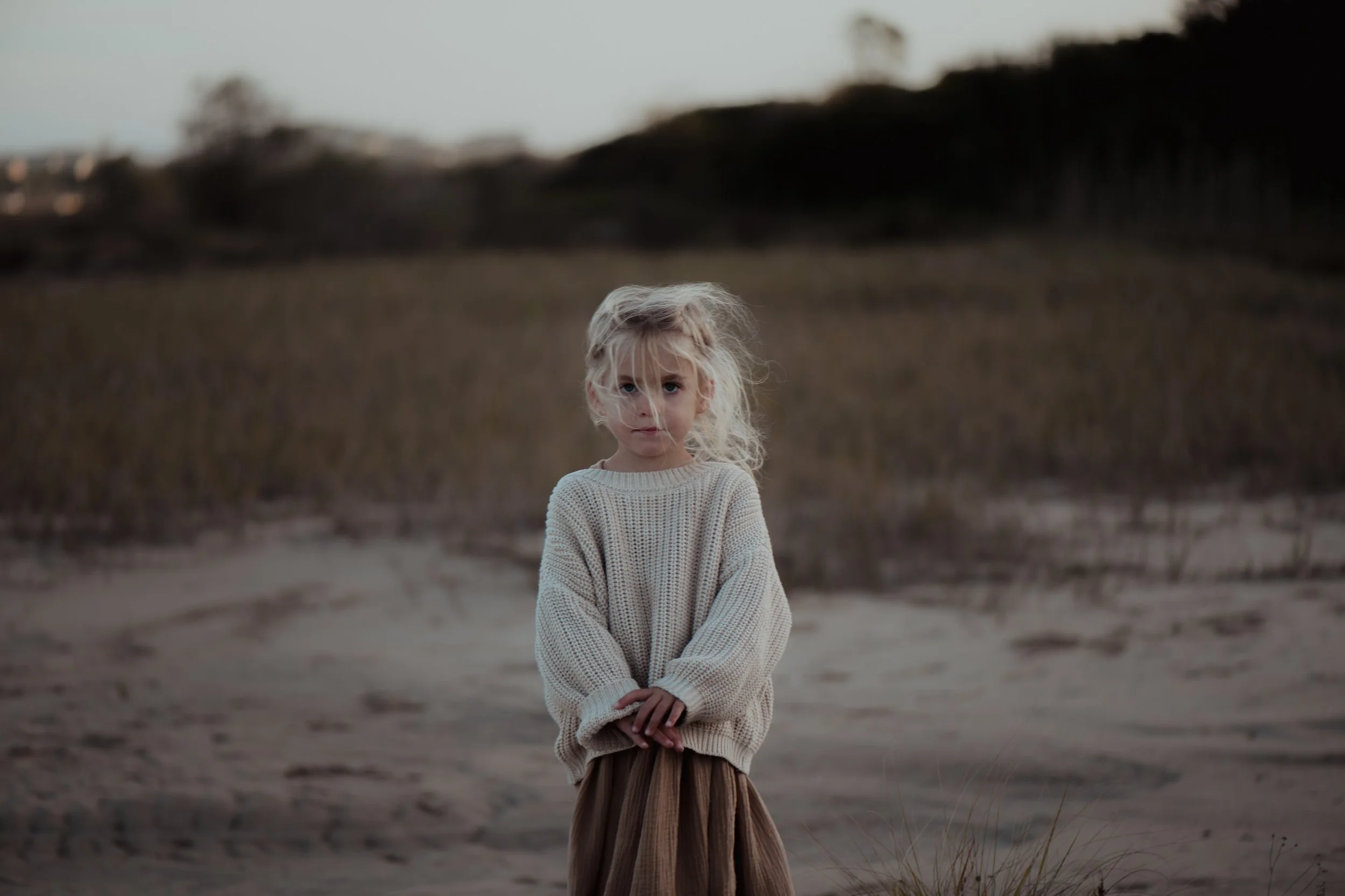 Ein junges Mädchen mit blondem, gewelltem Haar steht allein auf einer Sanddüne in einer offenen landschaftlichen Umgebung bei Sonnenuntergang.