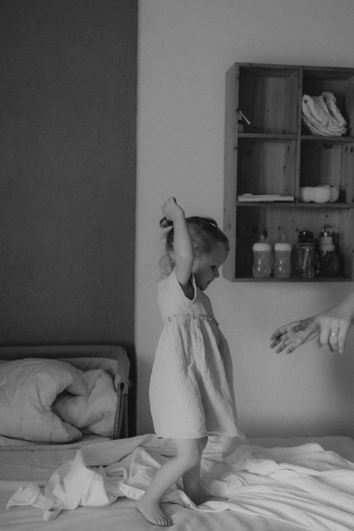 Ein junges Mädchen steht auf einem unaufgeräumten Bett und streckt die Arme nach oben. Es trägt ein ärmelloses Kleid. Im Hintergrund sind eine Wand, ein Wandregal mit verschiedenen Gegenständen und ein Teil eines Erwachsenen zu sehen, der sich mir de