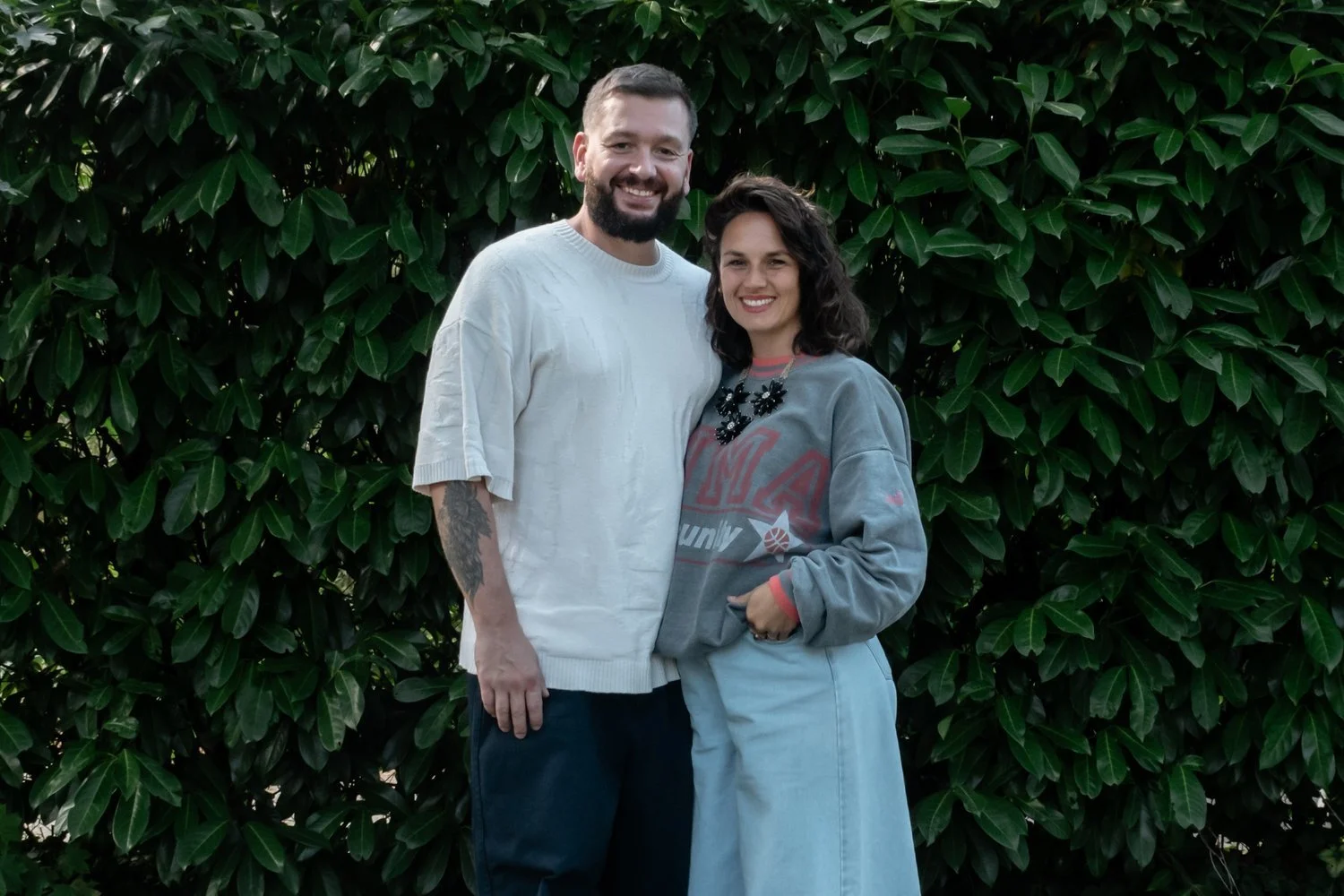 Un couple souriant posant devant un buisson vert dense.