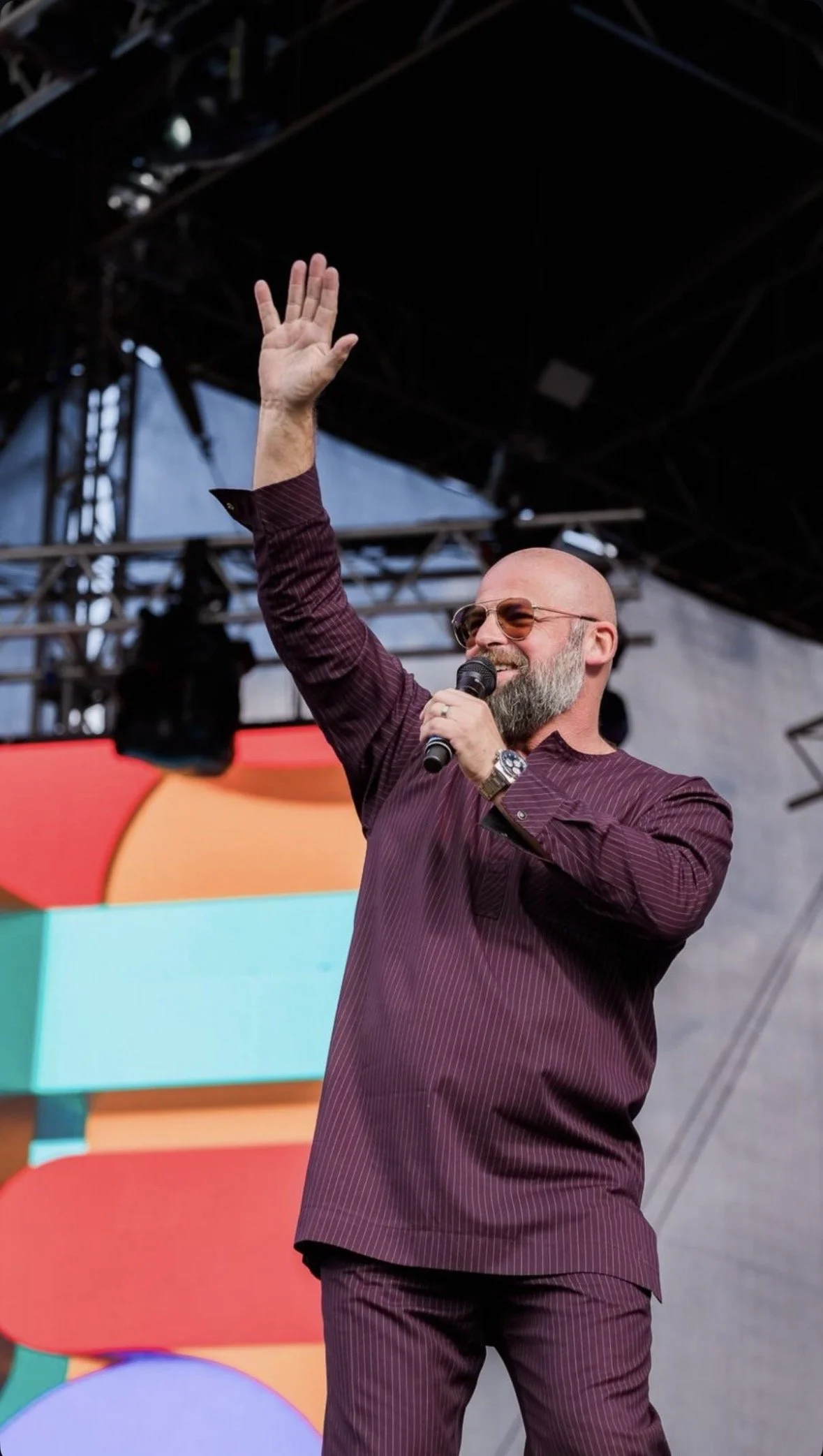 Un homme barbu portant des lunettes de soleil, une veste rouge à rayures et une montre, tient un microphone et fait signe avec la main lors d'un événement en plein air. En arrière-plan, un écran coloré et une structure de scène visible.