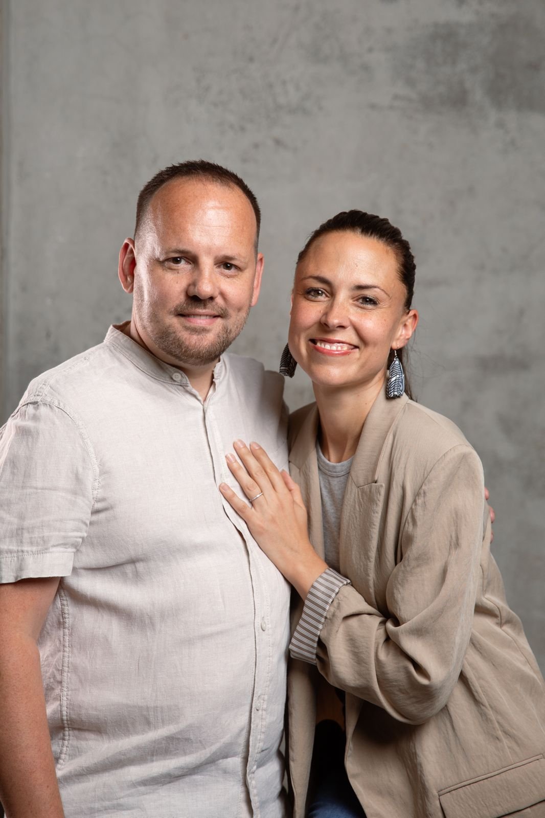 Un couple souriant posant devant un mur gris, l'homme porte une chemise beige et la femme une veste beige avec des boucles d'oreilles longues.