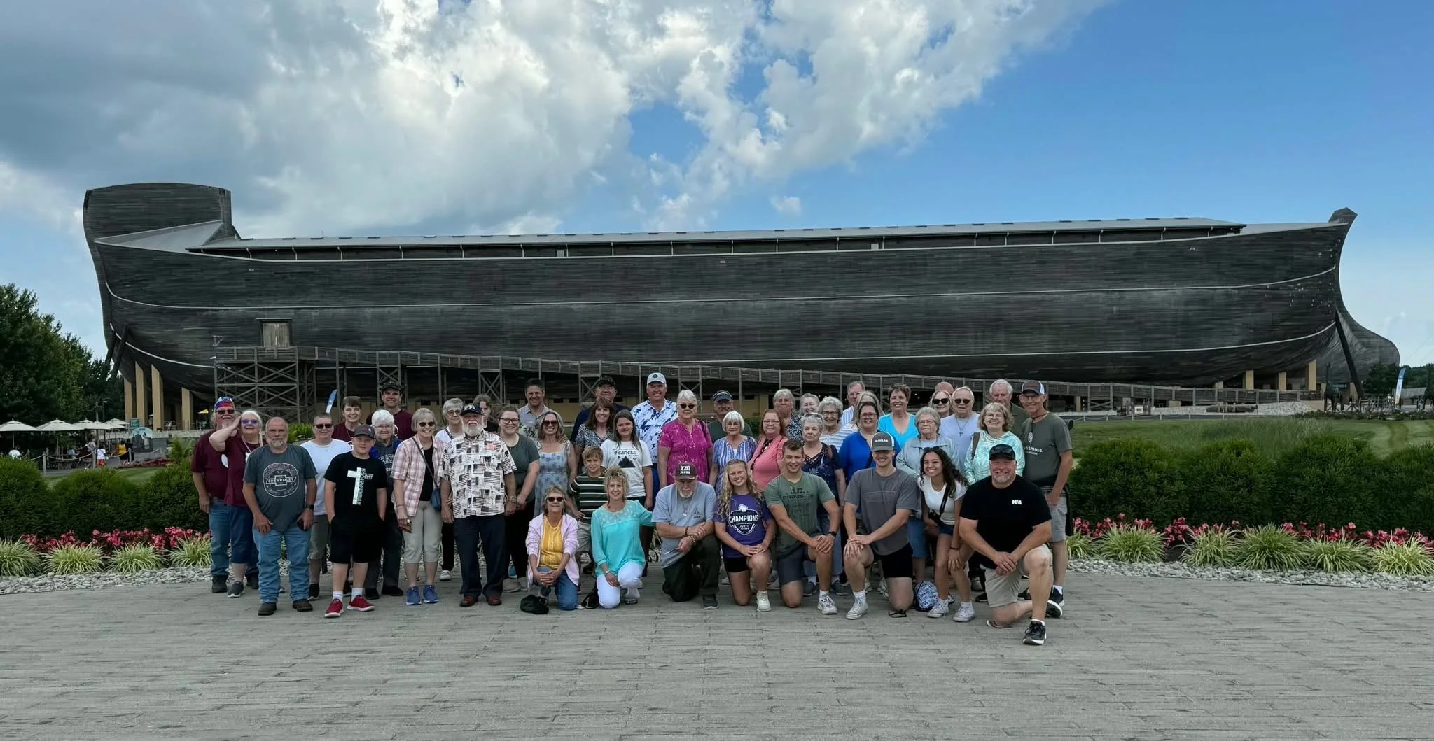 A group of people posing in front of the Ark Encounter, a replica of Noah's Ark at a bus trip taken by LaValle Advent Christian Church. The group includes children, adults, and seniors, with a partly cloudy sky overhead.