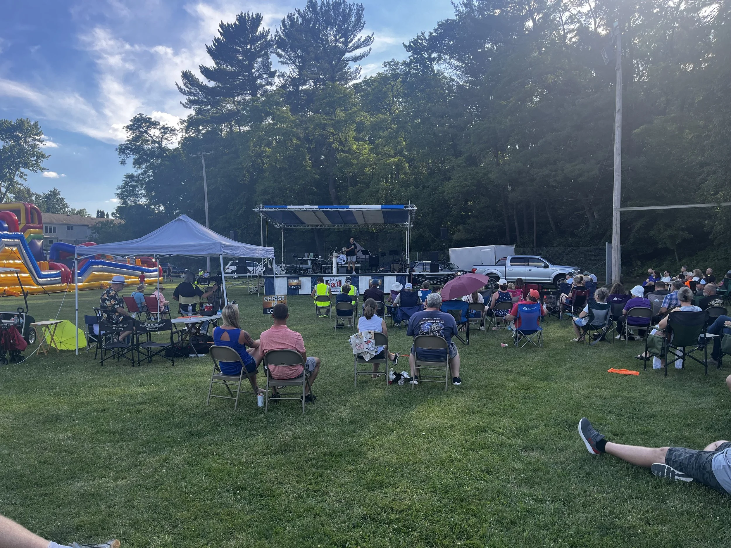 Praise in the Park Event for 150th Anniversary with a stage, audience sitting on chairs, tents, inflatable slides, and a background of tall trees and blue sky.