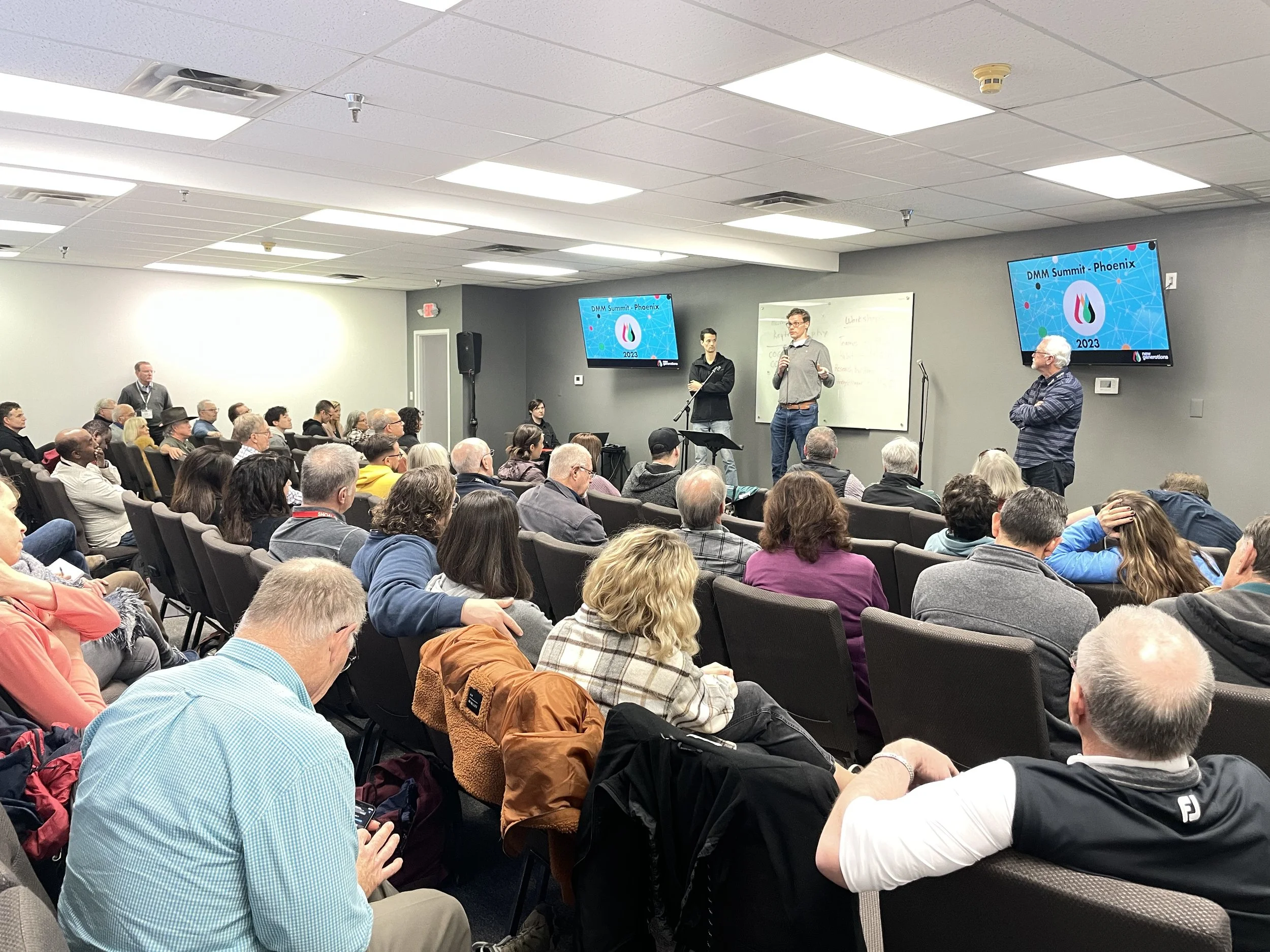 People attending a conference or seminar, sitting in chairs and listening to a speaker on stage in a conference room.