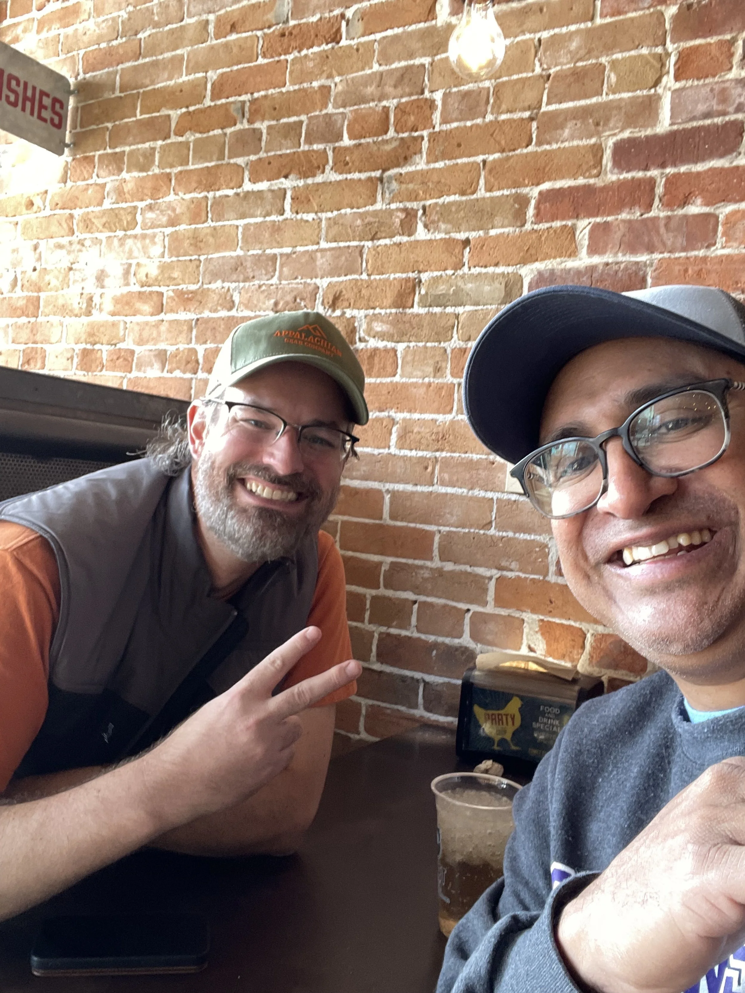 Two smiling men taking a selfie together at a restaurant with a brick wall background, one wearing glasses and a cap, holding up a peace sign, and the other wearing glasses and a baseball cap.