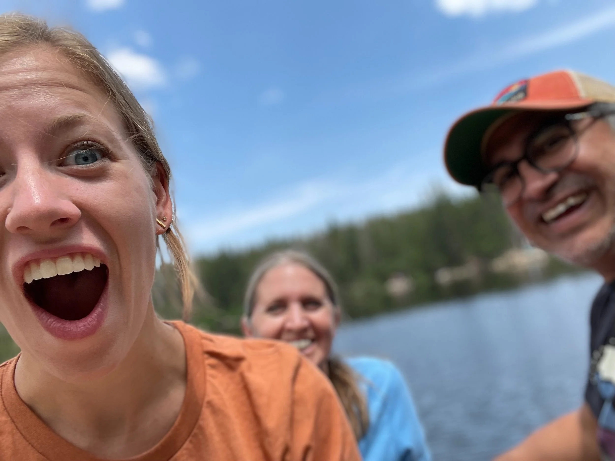 Close-up of three people smiling outdoors near a body of water with trees in the background. The person in the foreground has blonde hair, blue eyes, and is wearing an orange shirt. The person on the right is wearing glasses, a baseball cap, and a dark shirt. The person in the middle has long hair and is wearing a blue shirt.