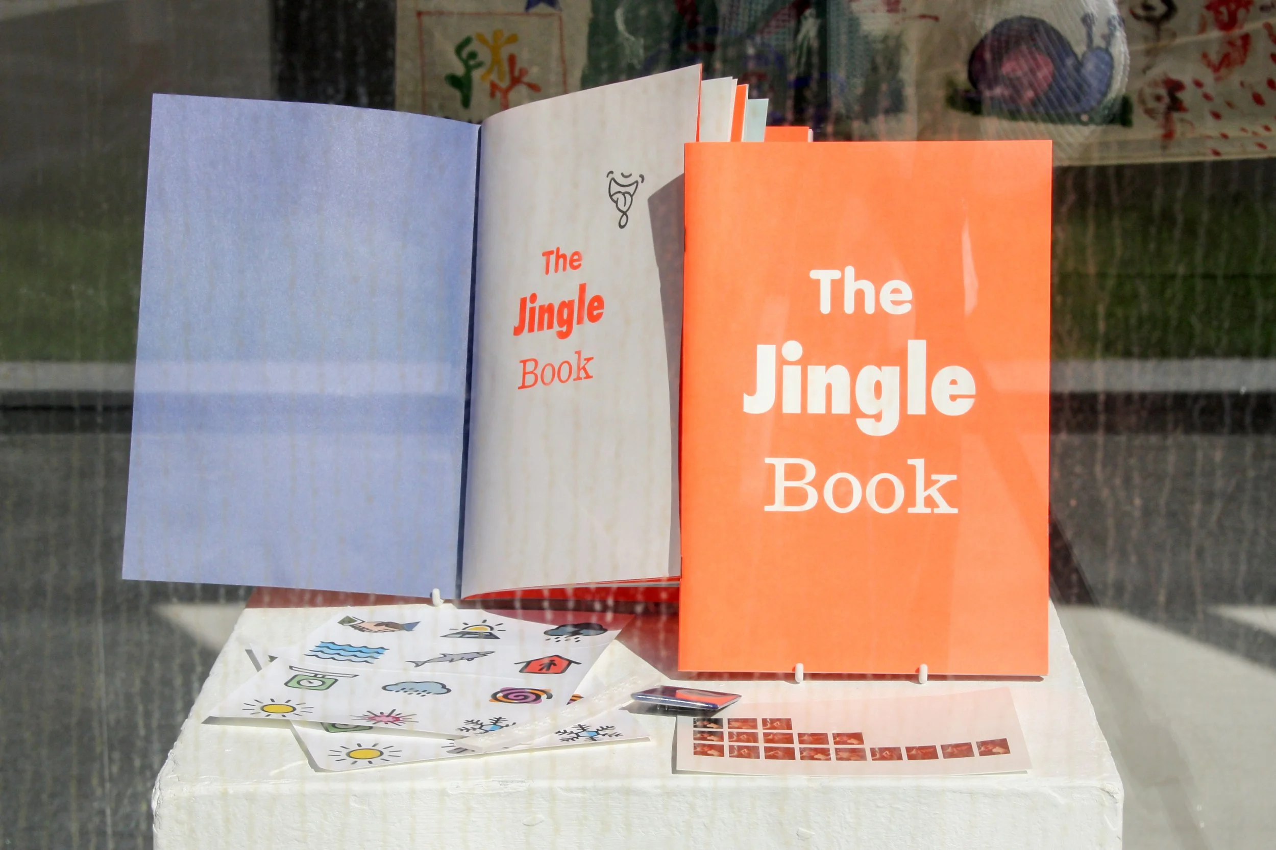 Exhibition photograph of one portrait-orientated book, slightly larger than A5, with a fluorescent orange cover and the title ‘The Jingle Book’ across three lines, each word centred in large white letters and each word in a different font, alongside