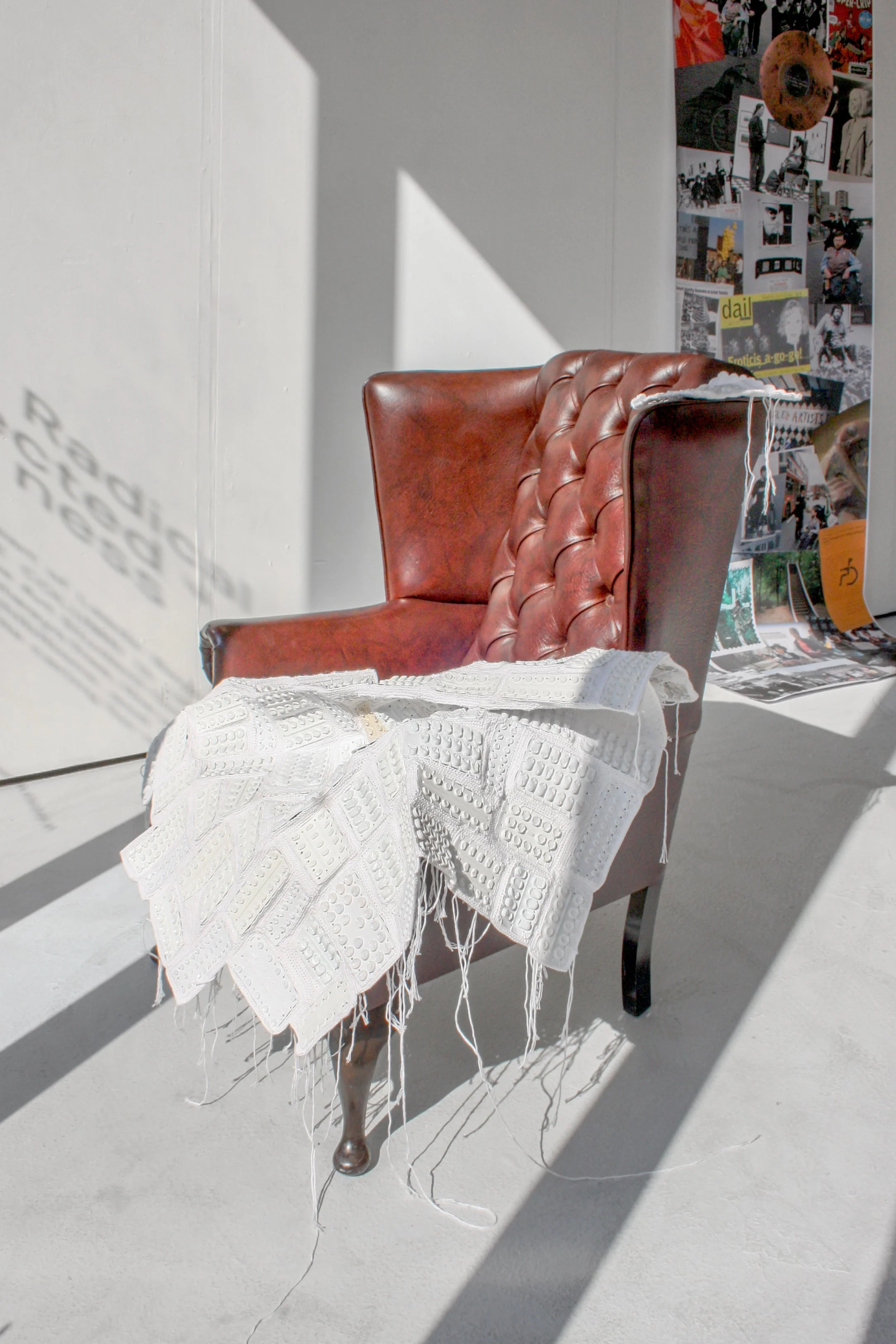 Exhibition photograph of ‘Memento Mori’ a mixed-media sculpture made up from empty and used white pill packets and white cotton, displayed on a vintage brown leather armchair. The sculpture is shown with the white vacuum-formed raised pill packets to
