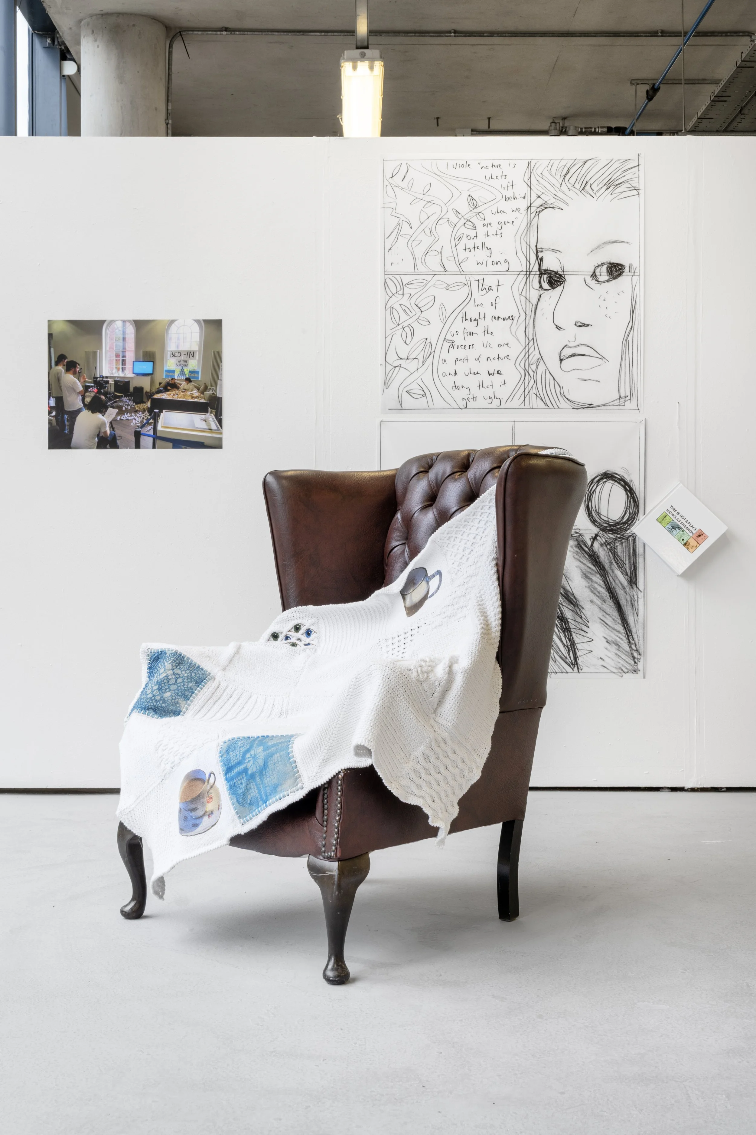 Exhibition photograph of ‘Idle Hands’ a mixed-media textile blanket made up of white cotton, embroidery thread, three calico cyanotyped squares. The blanket is displayed on a vintage brown leather armchair, draped over its arm rest and coming to rest