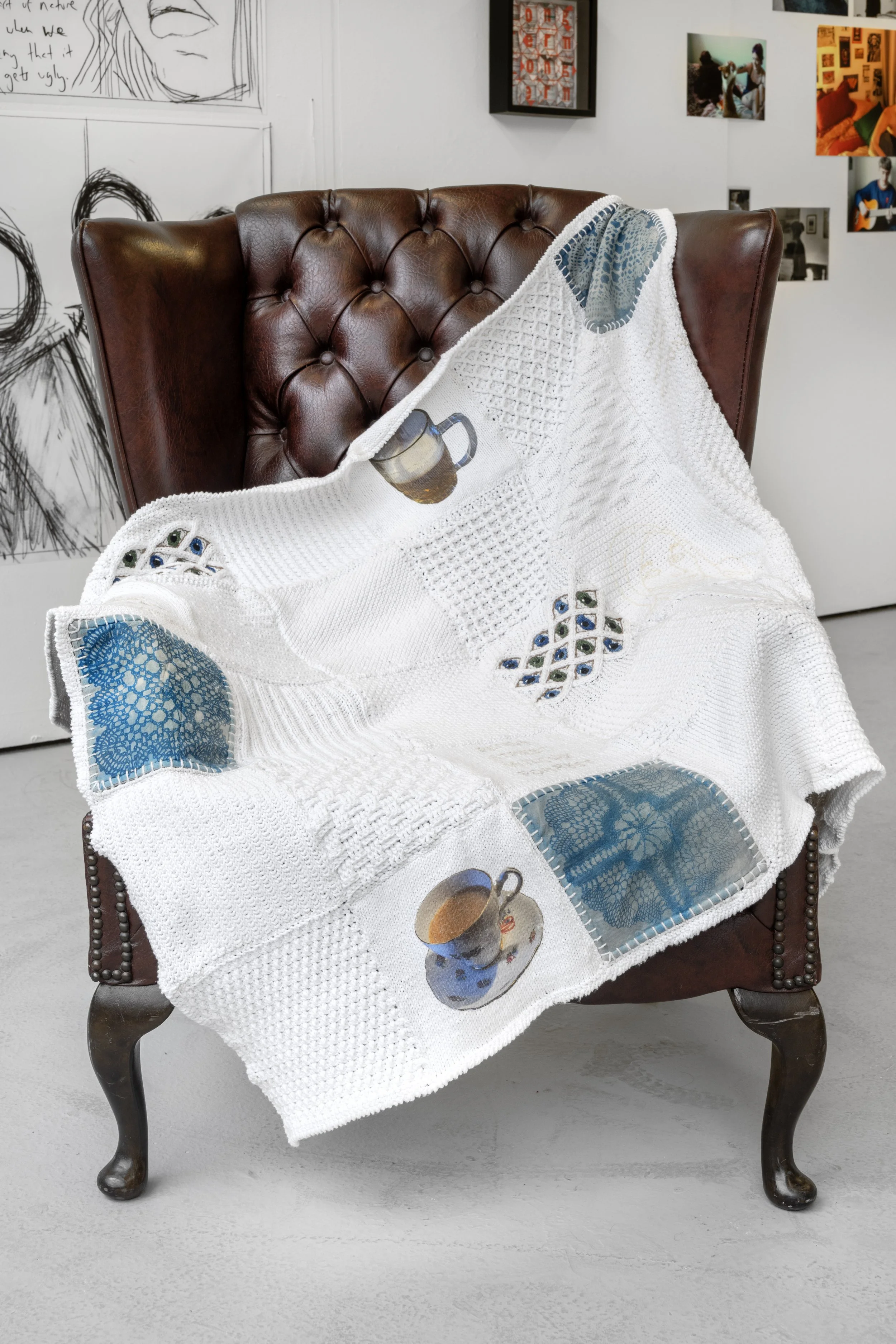 Exhibition photograph of Joanne Tiffany's ‘Idle Hands’ a mixed-media textile blanket made up of white cotton, embroidery thread, two calico cyanotyped squares. The blanket is displayed on a vintage brown leather armchair, draped over its arm rest and