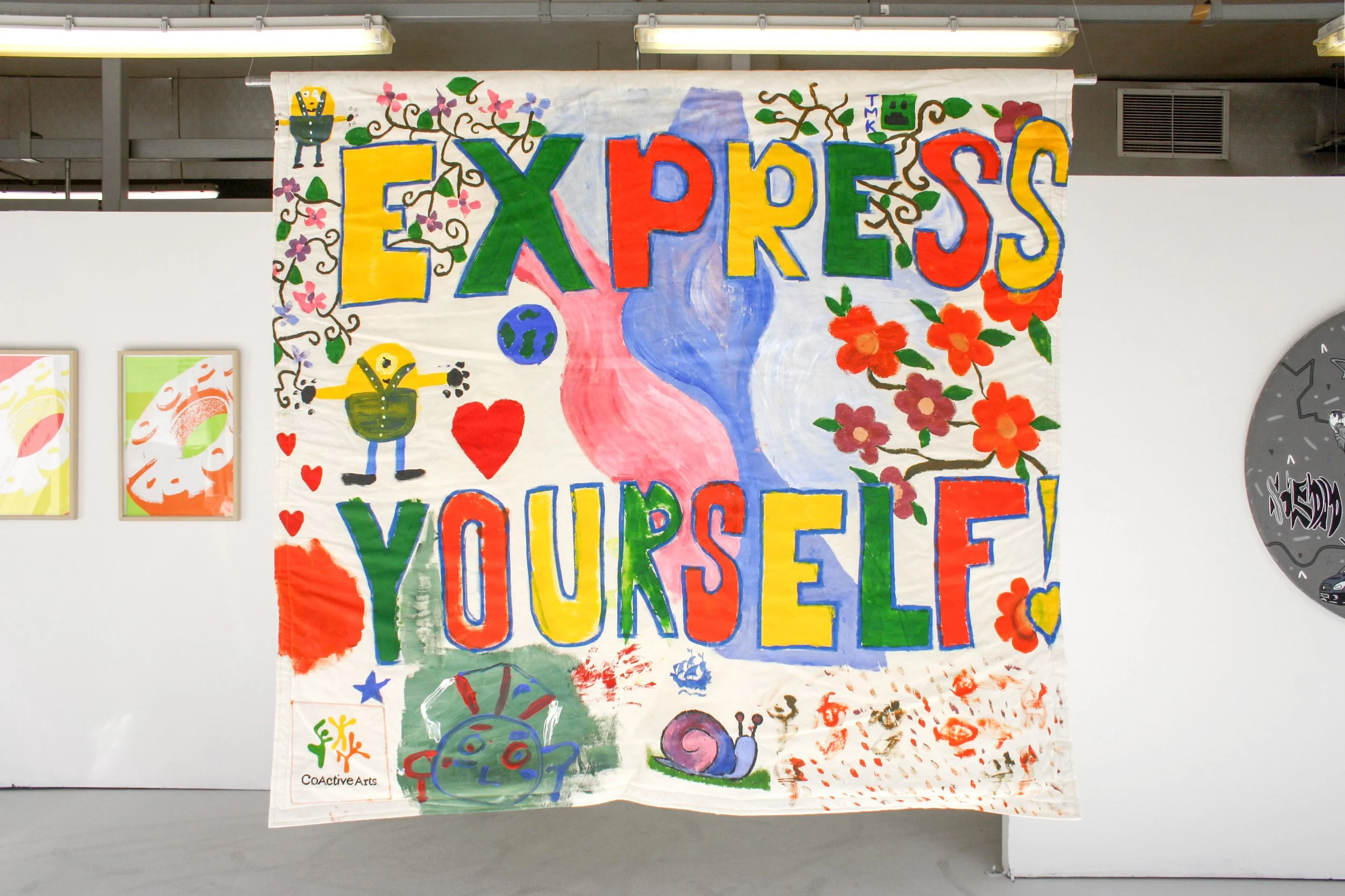 Exhibition photograph of the front view of a collaborative artwork banner of 58x53 inches with the words ‘Express Yourself’ in alternating colours of yellow, green, and orange. The background is interspersed with pink, dark and light blue pattern, a