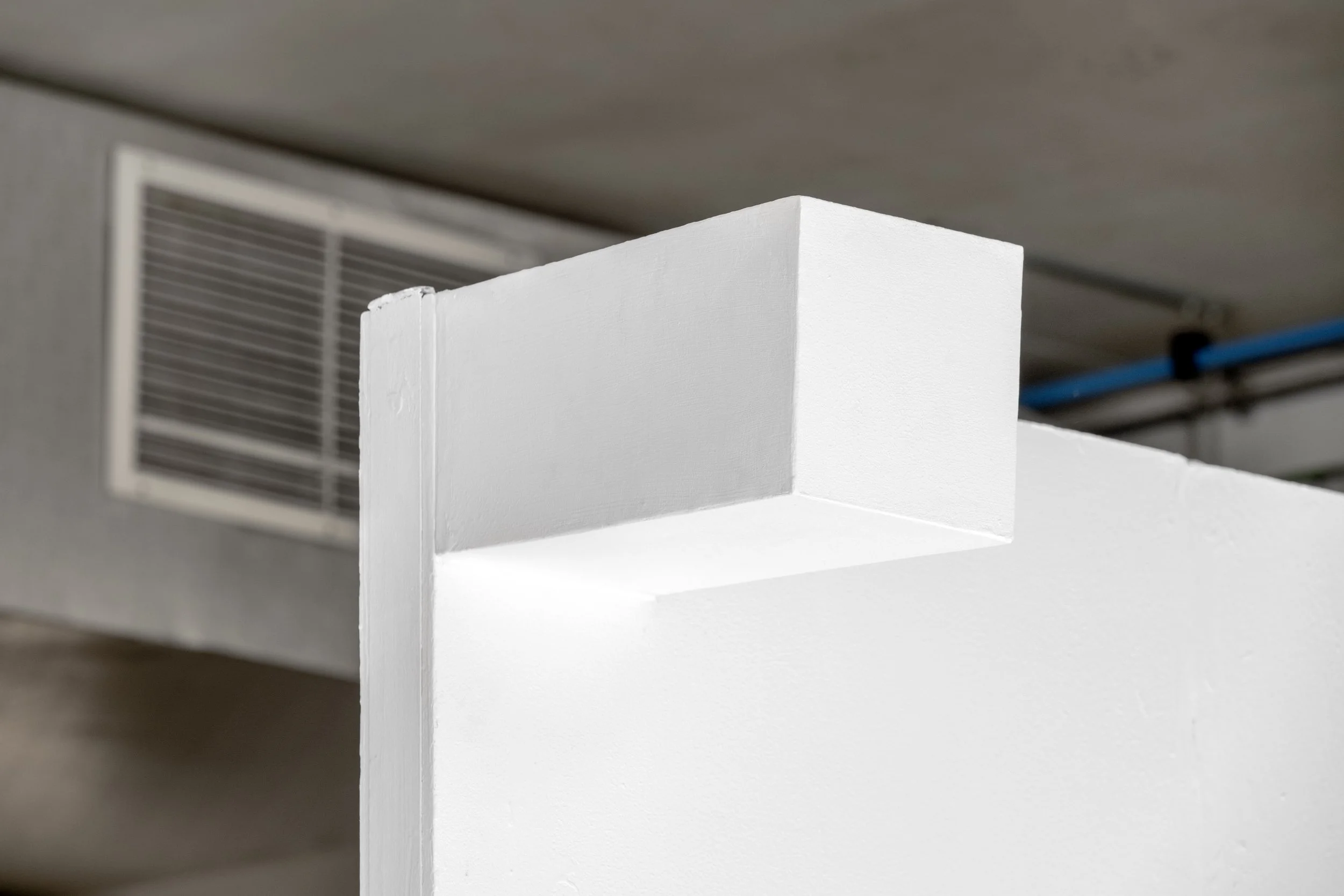 Harry Meadley's artwork photographed high up on a white wall in the White Column space. A white shoe box sized form protrudes from the upper part of a white gallery wall. It is positioned so even a tall person would look up at it. The box’s surface i