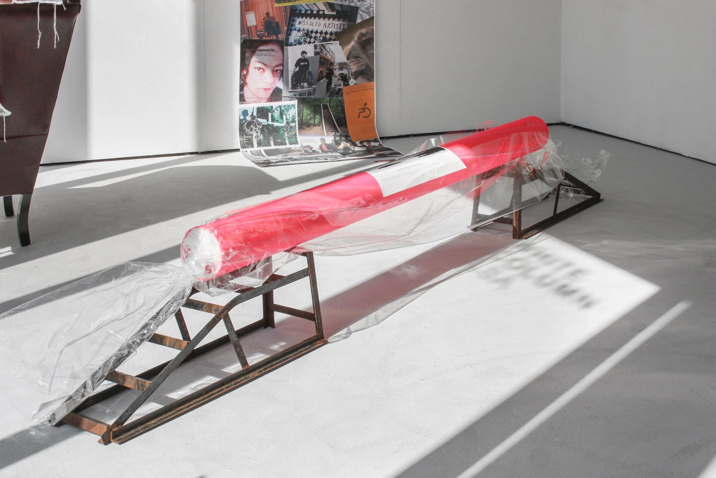 Exhibition photo of ‘Rock#1’ a 240cm-long tubular mixed-media sculpture resembling a stick of rock, displayed on two worn old rusty black metal car ramps, shown on a dove grey floor, surrounded by other artworks. Photograph credit: Harry Meadley.