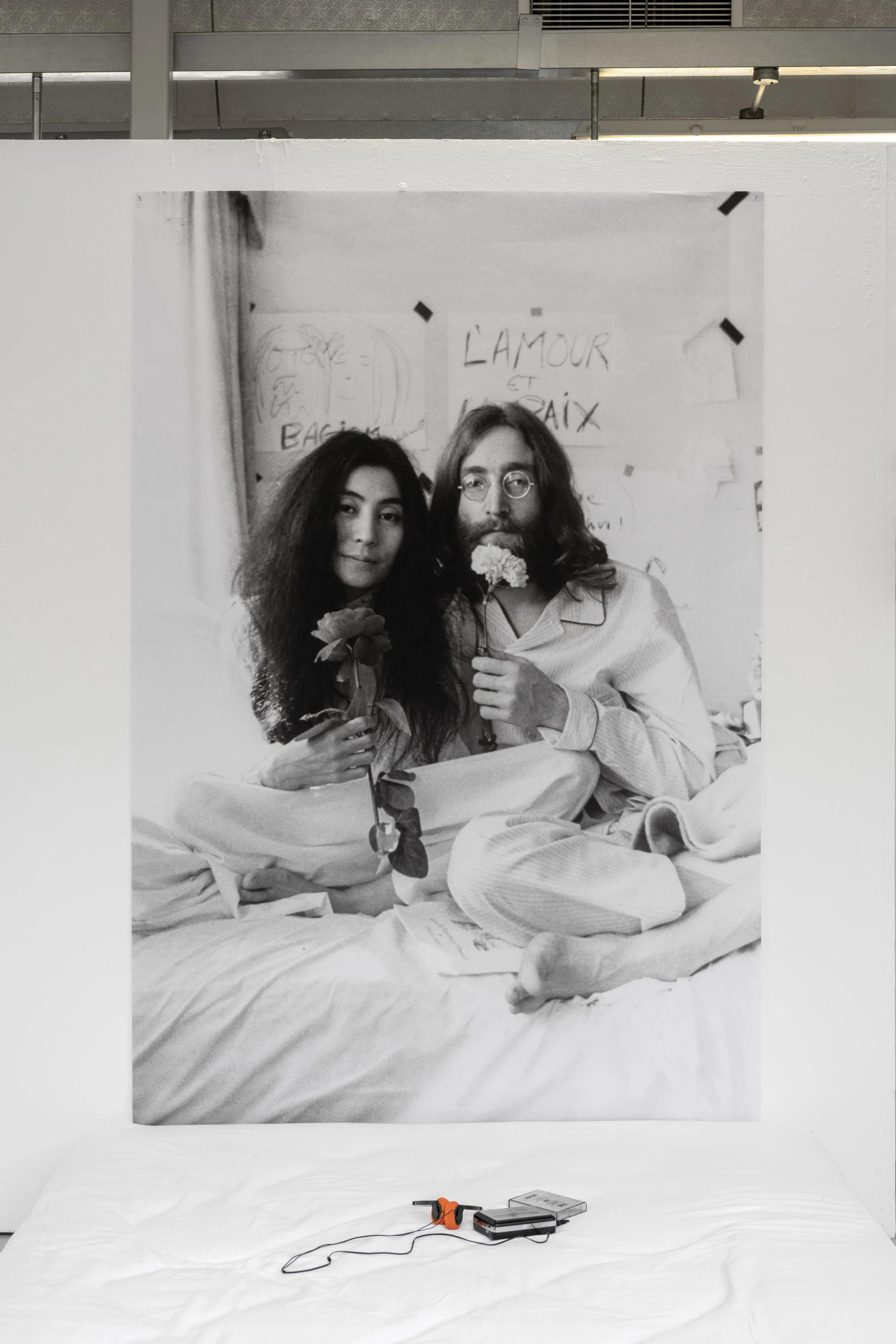 Exhibition photograph of  Yoko Ono's 'Bed-In' a black and white photograph against a white wall in the White Column space. The artwork photograph has been taken of the performance ‘Bed In’ of artist Yoko Ono (Left) and John Lennon (right) sitting on 
