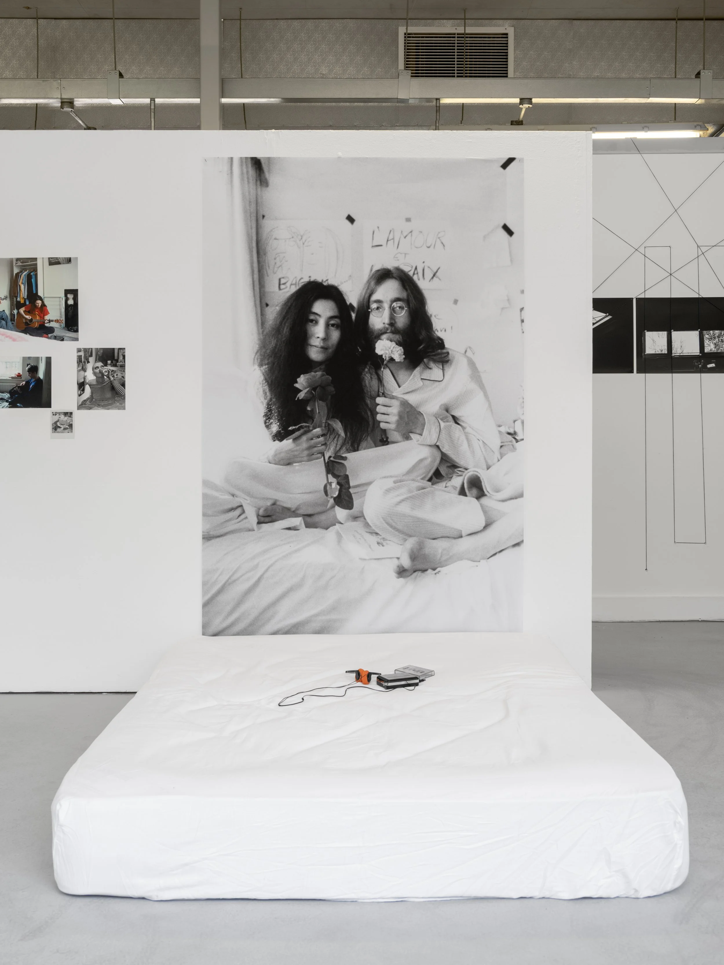 Exhibition photograph of Yoko Ono's 'Bed-In' a black and white photograph against a white wall in the White Column space. The artwork photograph has been taken of the performance ‘Bed In’ of artist Yoko Ono (Left) and John Lennon (right) sitting on a