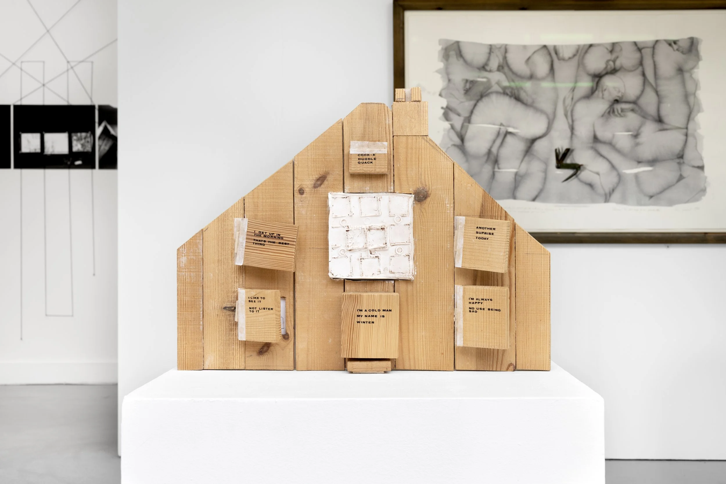 Exhibition photo of Vivify Project's ‘Wooden Dream Houses’ displayed on a white plinth placed on a grey floor within the White Column space. The sculpture depicts the front of a house, made from rough grained timber, jig sawed pieced together leaving
